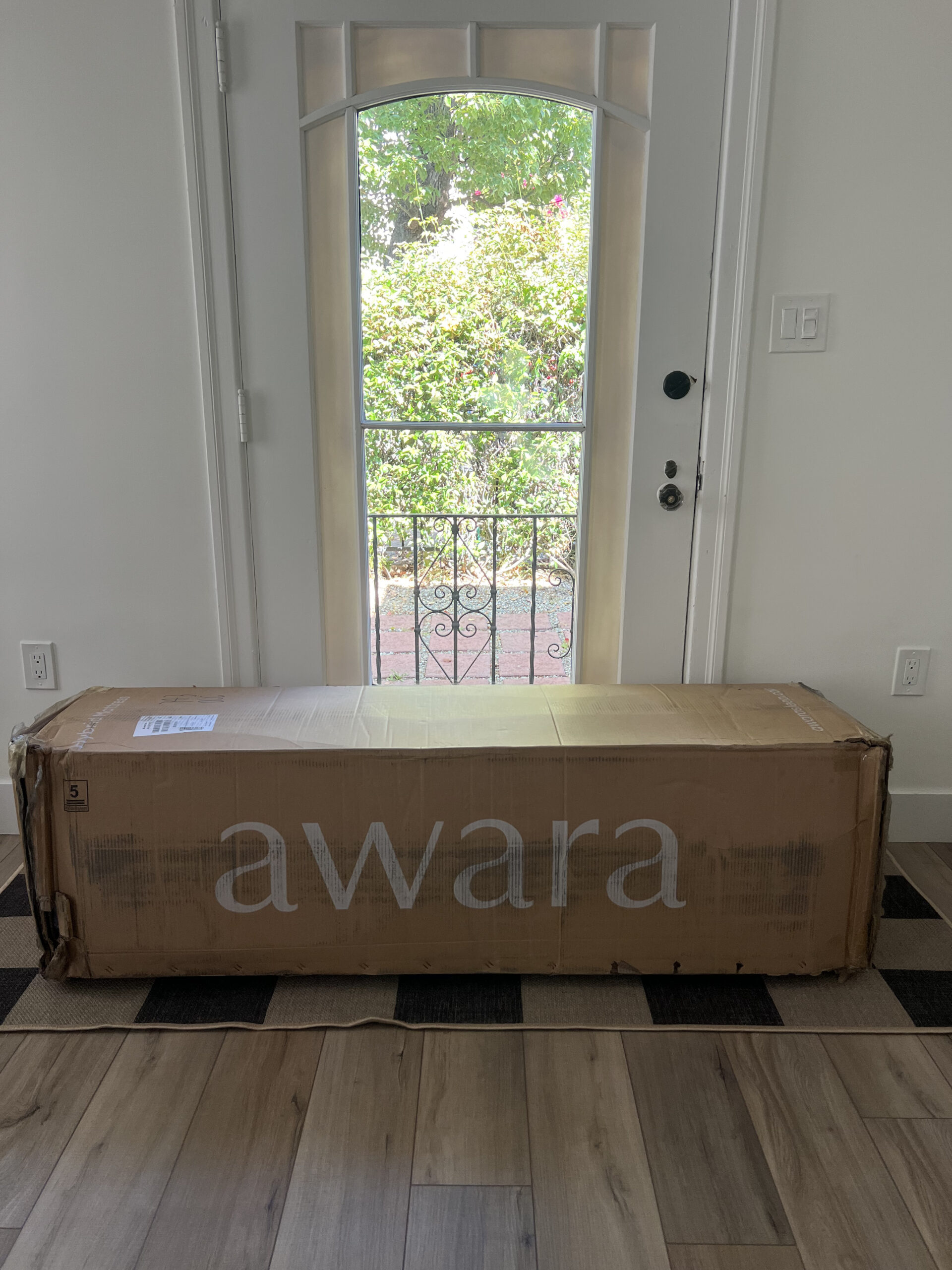 A large cardboard box labeled "Awara" is placed on the floor in front of a glass door with a view of a garden outside.