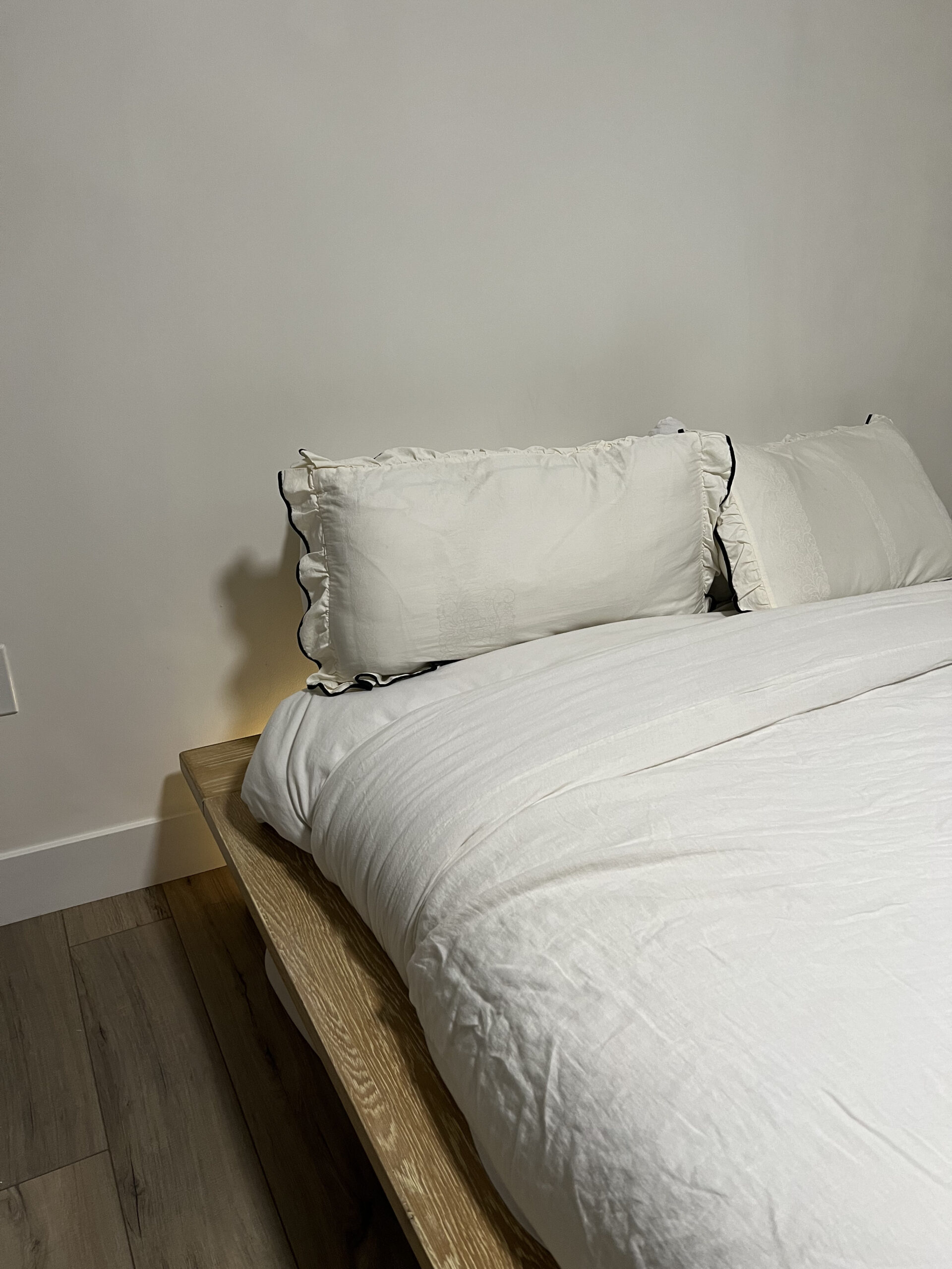 A neatly made bed with white pillows and a white comforter on a wooden bed frame, set against a plain white wall. The floor is wooden.