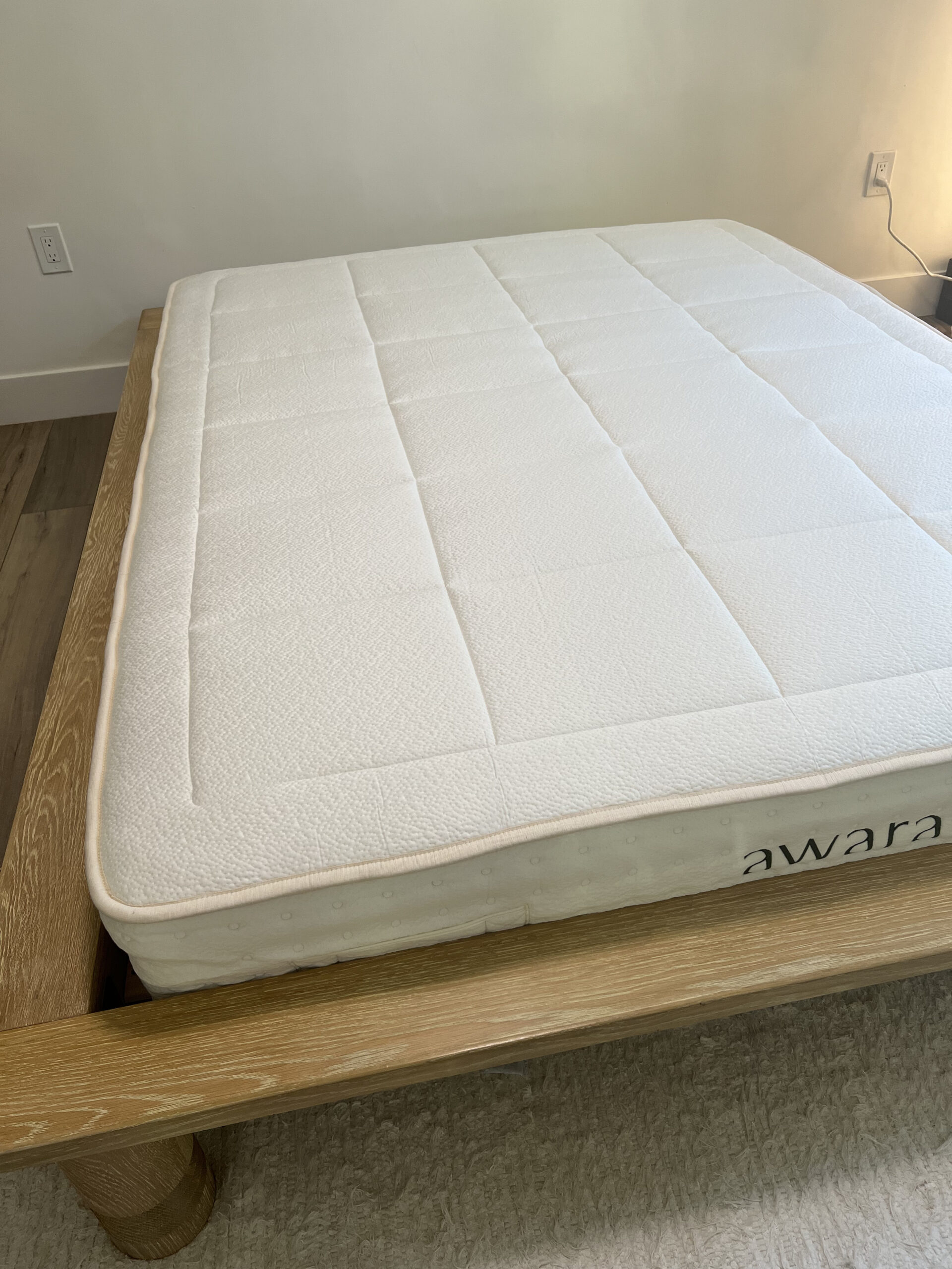 A white, quilted mattress labeled "Awara" placed on a wooden bed frame in a minimalist, well-lit room. An electrical outlet is visible on the wall.