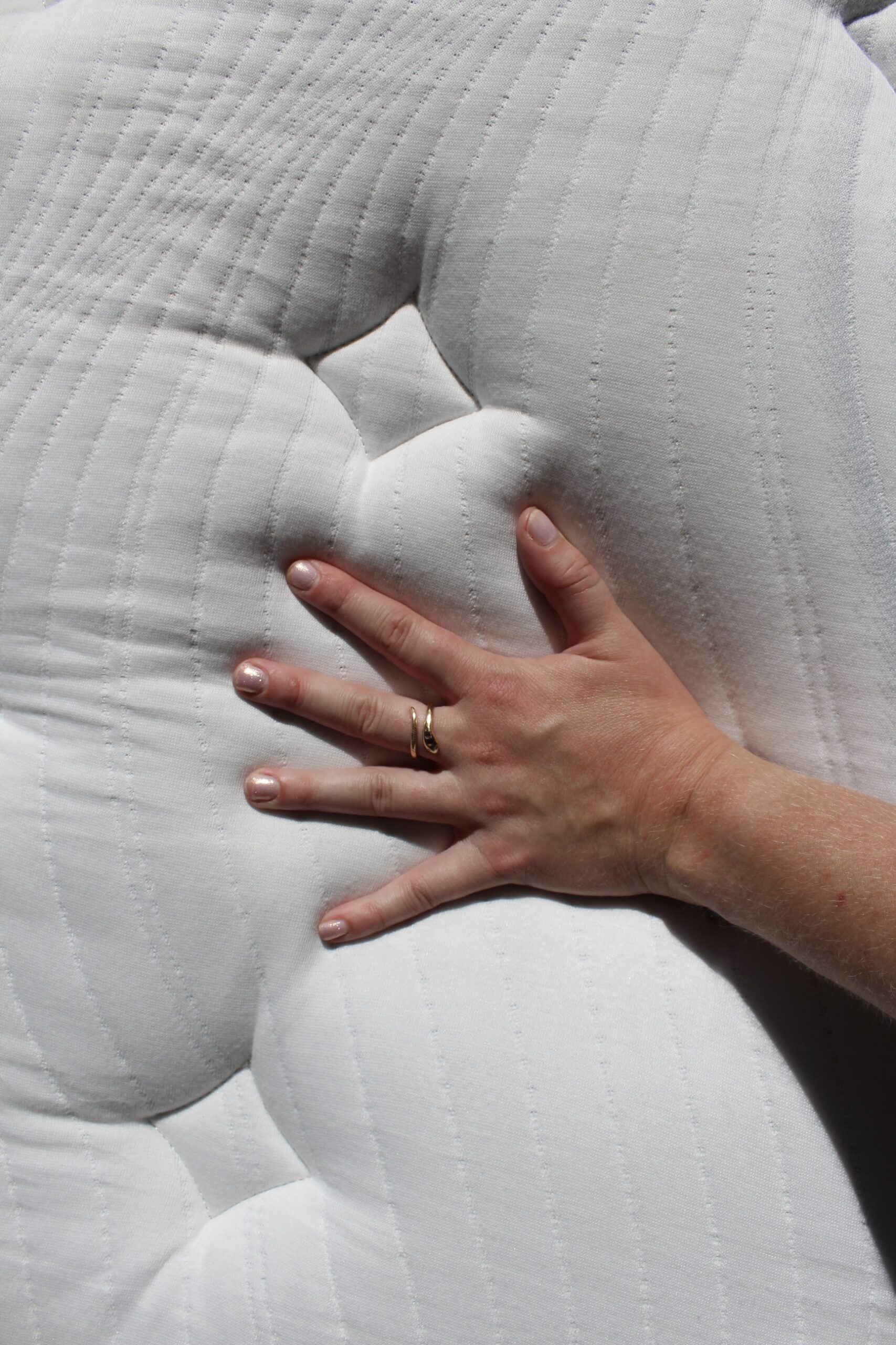 A hand with a wedding ring presses down on a white, tufted mattress to test its firmness.