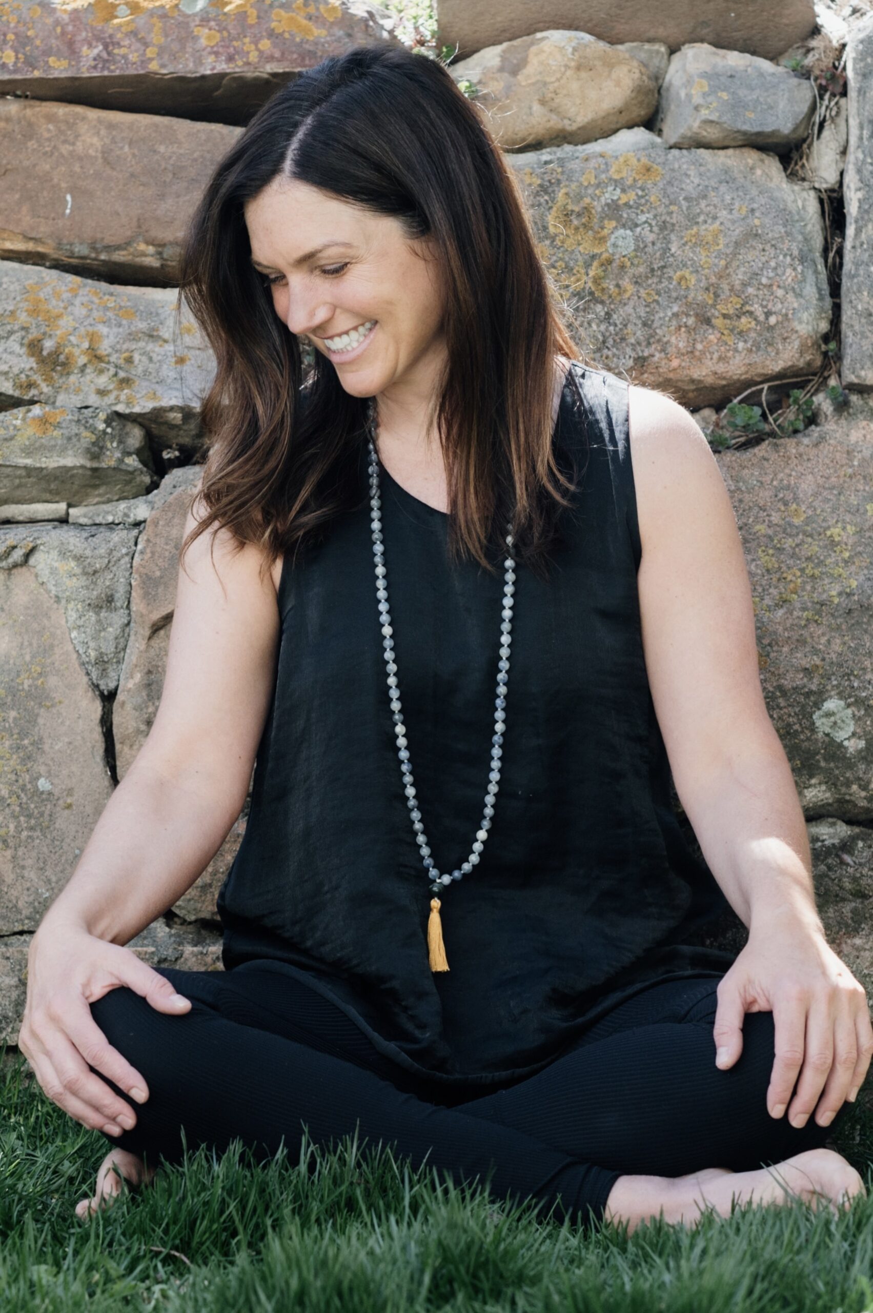 Woman smiling with brown hair seated cross-legged on the grass with a stone wall behind them, wearing a beaded mala necklace that is blue with a gold tassel.