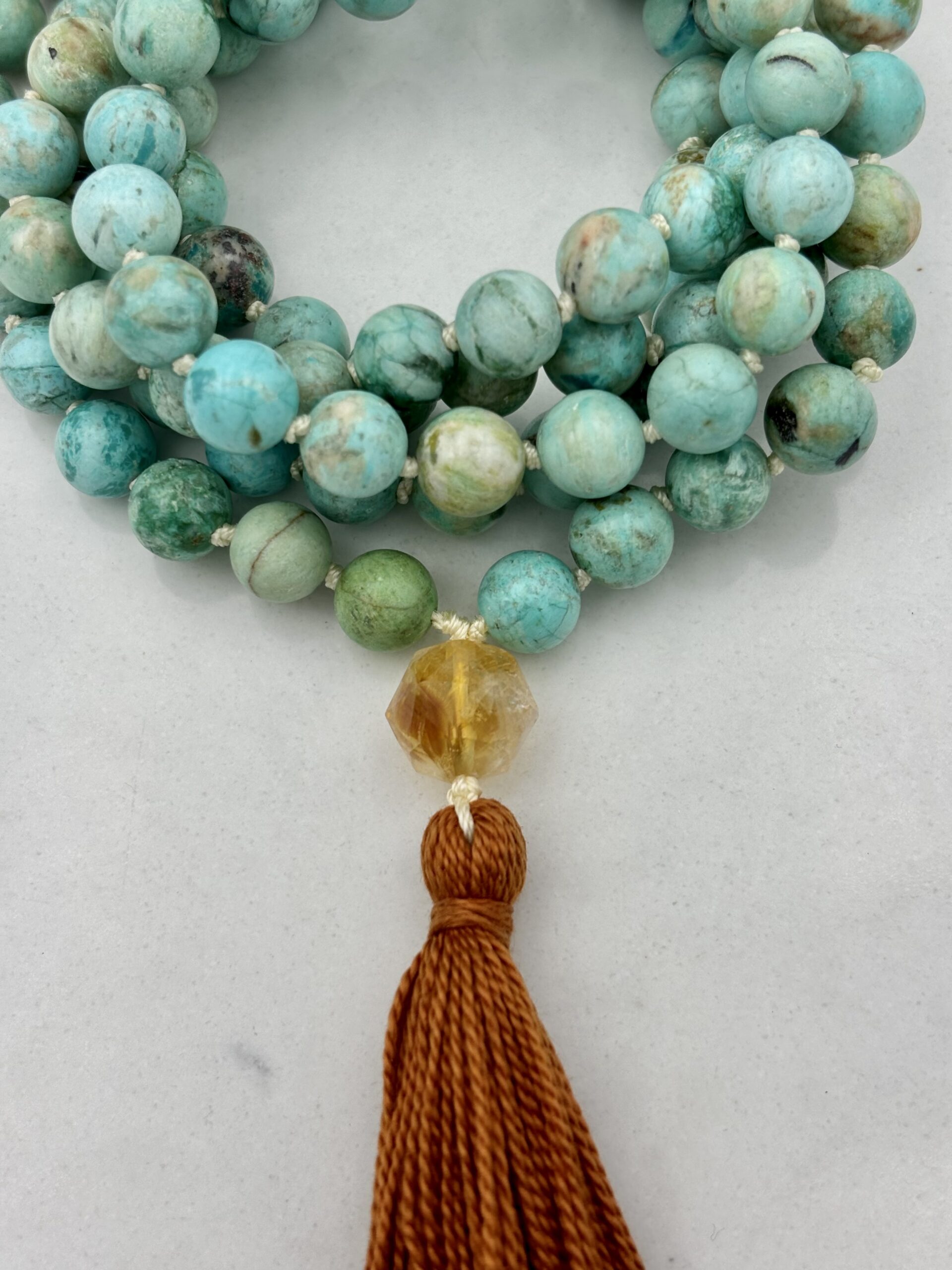 A beaded mala in a turquoise color with a brown crystal and brown tassel attached.