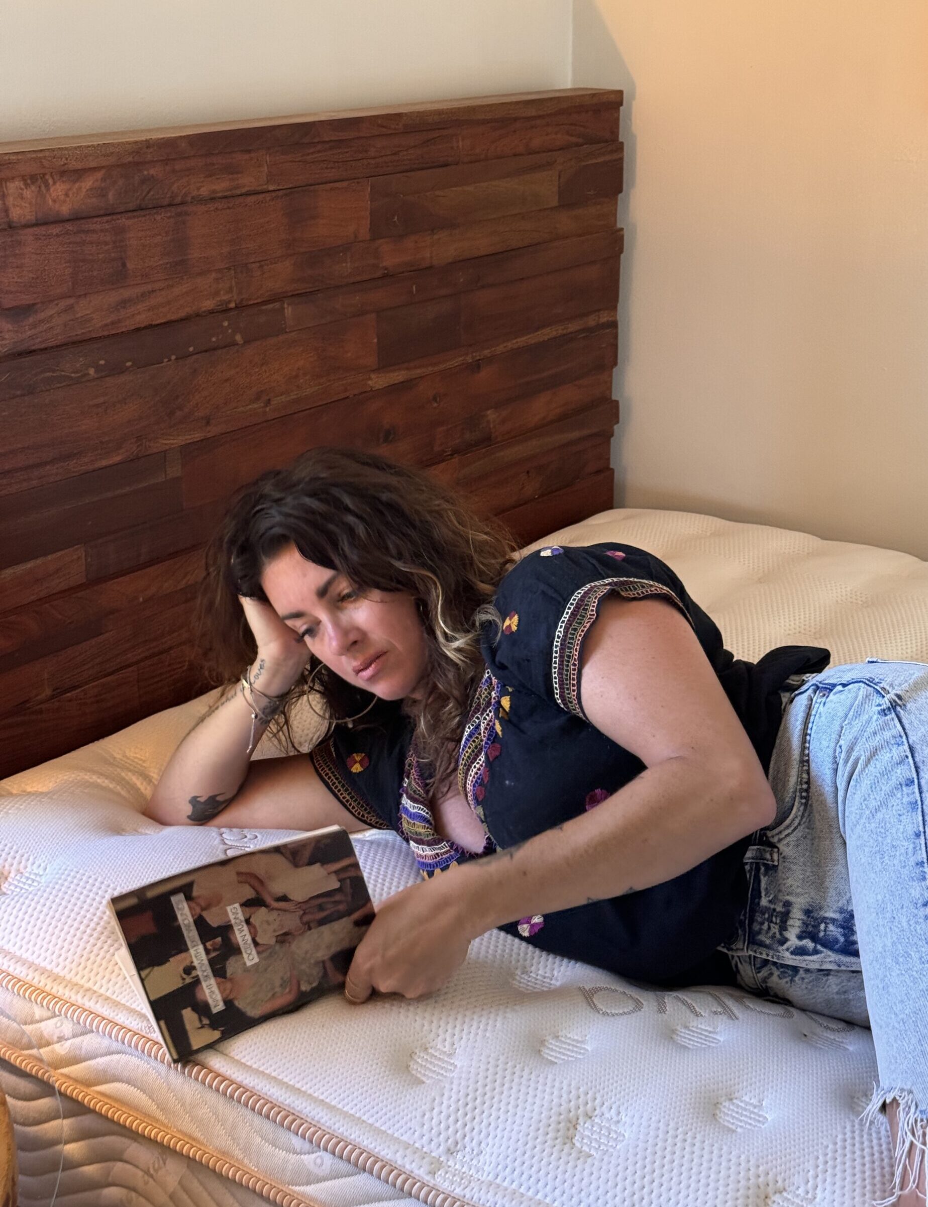 A person with wavy hair is lying on a bed, wearing a dark shirt and jeans, reading a book.