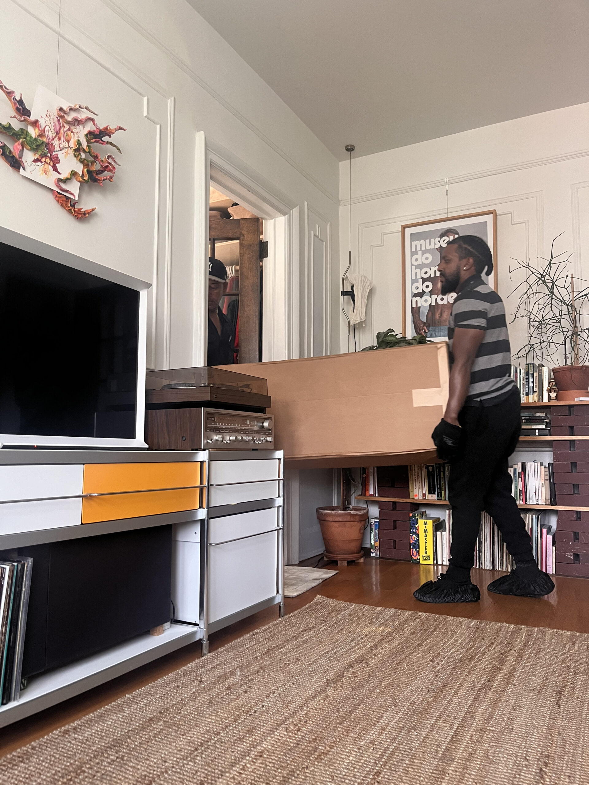 A person wearing a striped shirt and dark pants is carrying a large cardboard box inside a room with shelves, books, a TV, and decor.