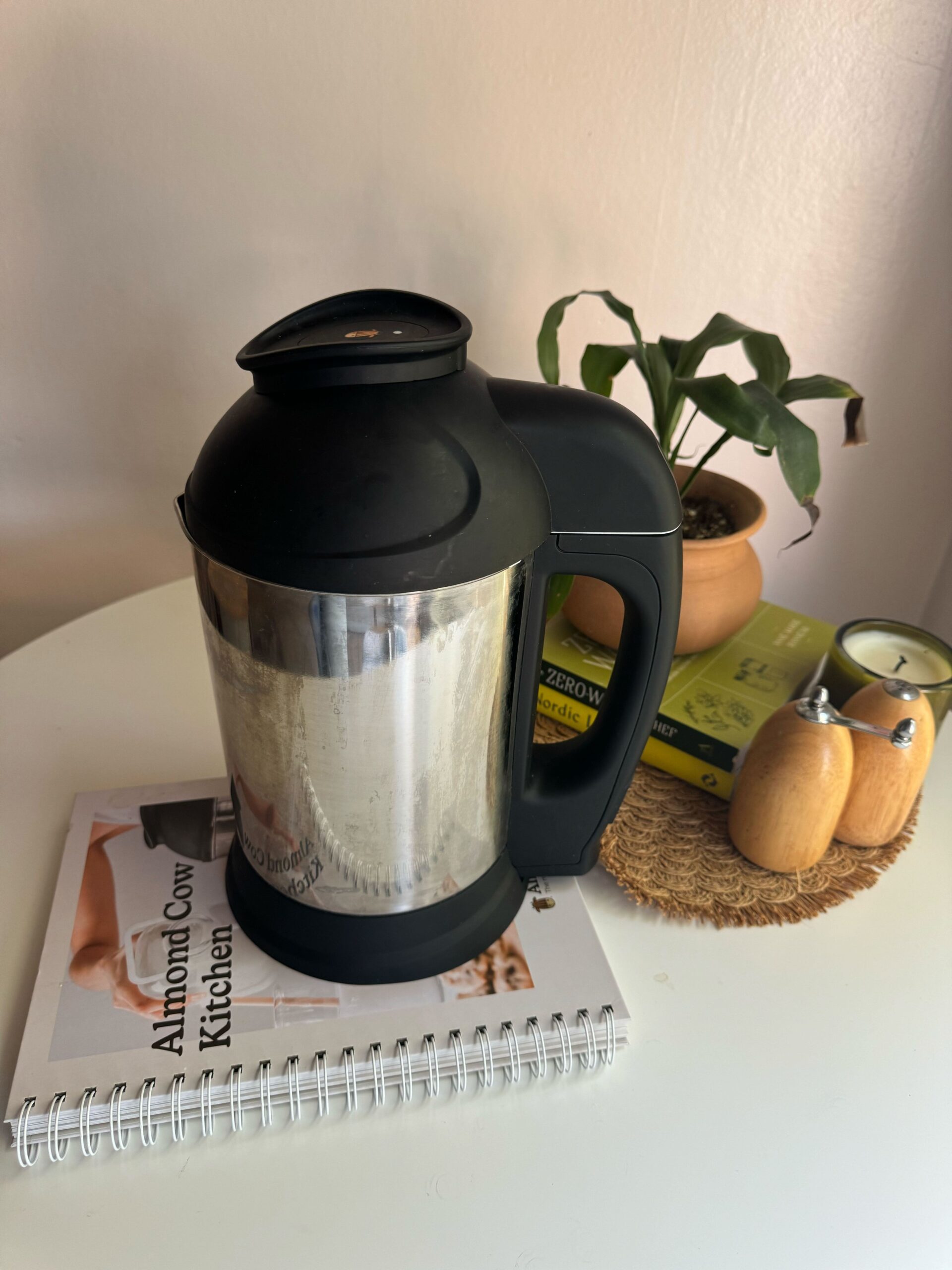 A stainless steel and black Almond Cow milk maker is placed on a table, alongside a notebook, potted plant, candles, and salt and pepper shakers.