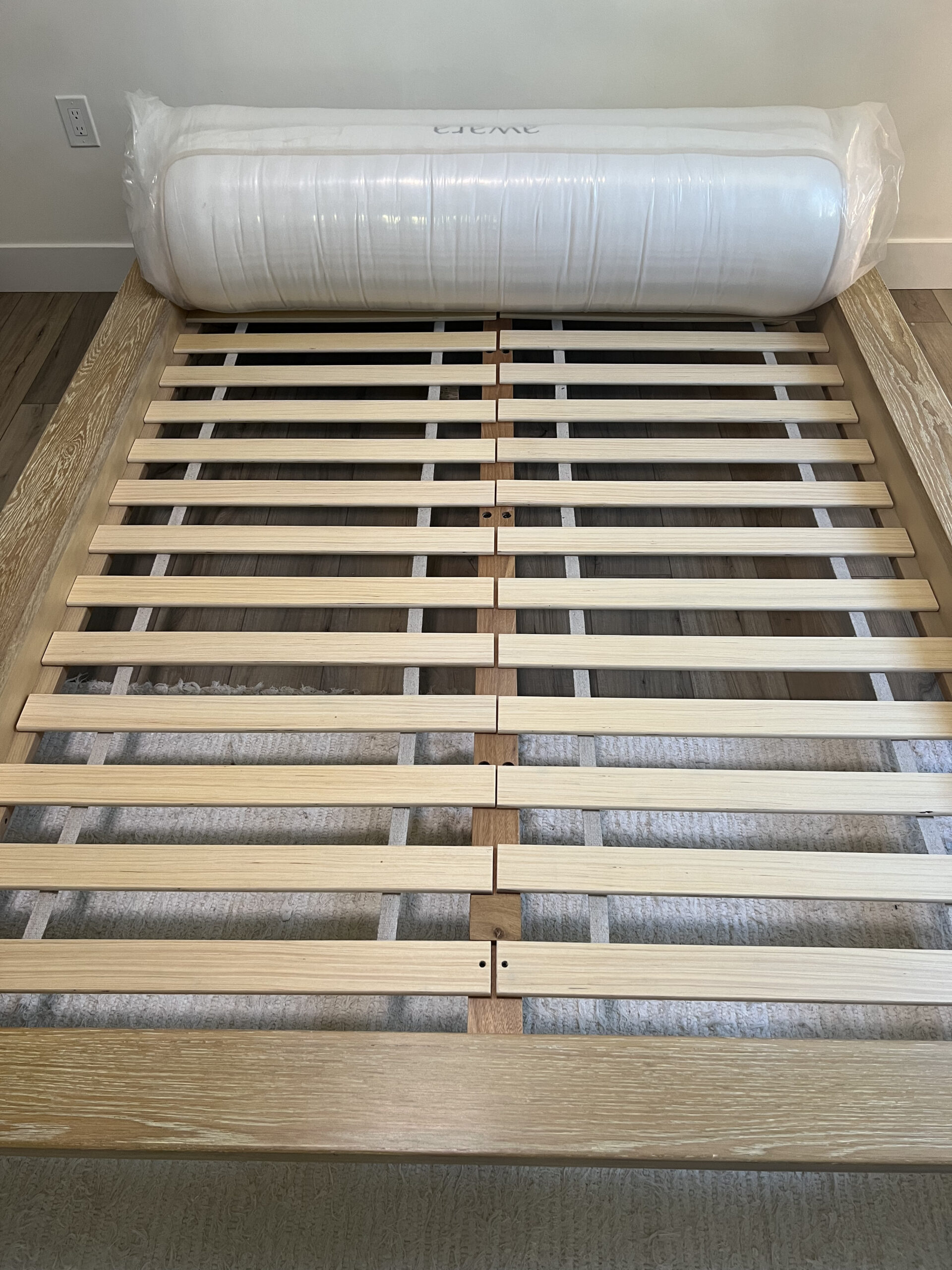 A wooden bed frame with evenly spaced slats is shown with a rolled-up mattress in plastic packaging placed on top.