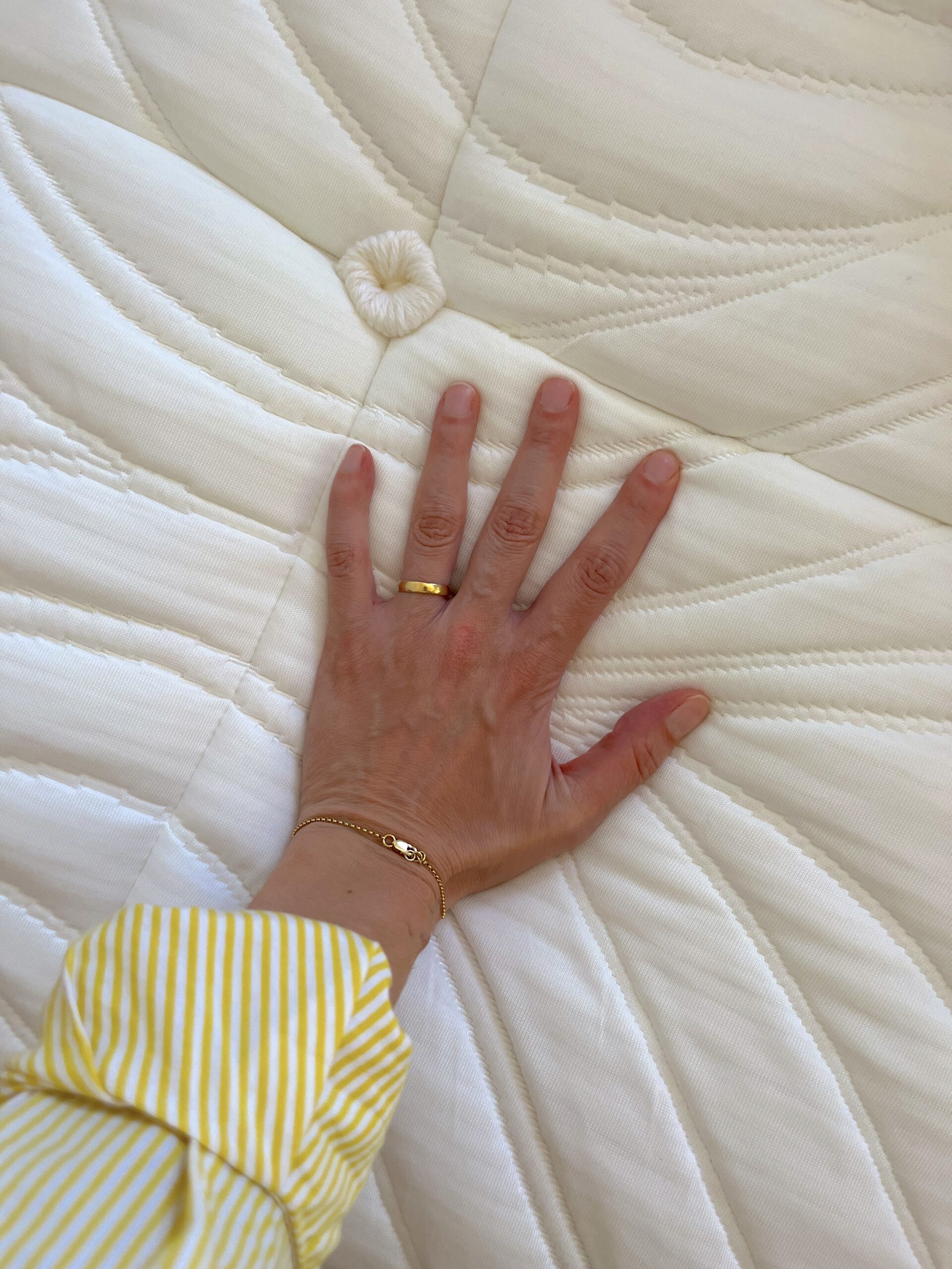 A hand adorned with gold rings and a bracelet rests on a white, textured organic mattress. The person is wearing a yellow and white striped shirt.