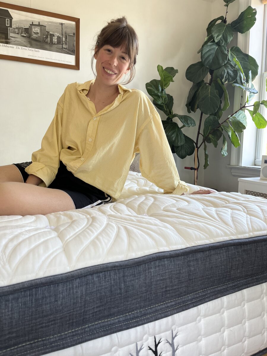 A person with a yellow shirt and black shorts sits on a bed with a white and gray mattress in a well-lit room with a plant and framed picture on the wall.