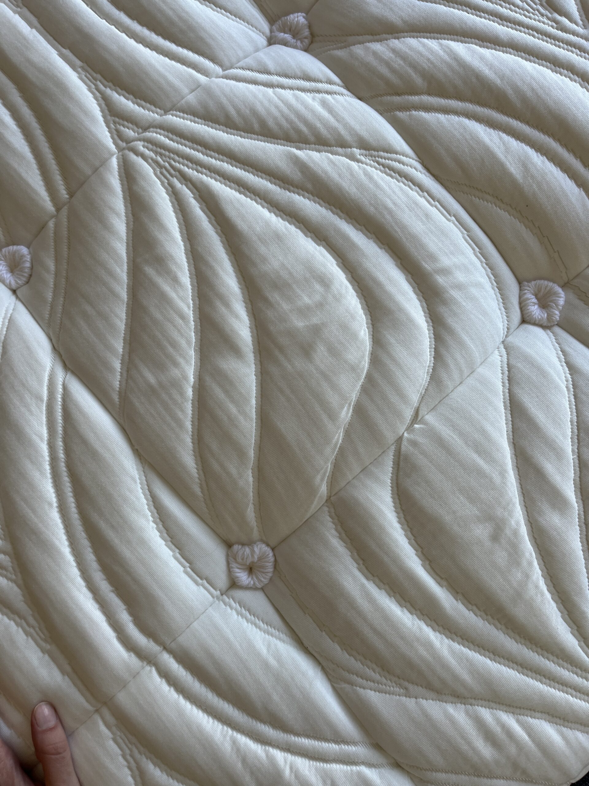 Close-up of a quilted mattress with a pale cream-colored fabric and decorative stitched patterns. A hand partially appears at the bottom left corner.