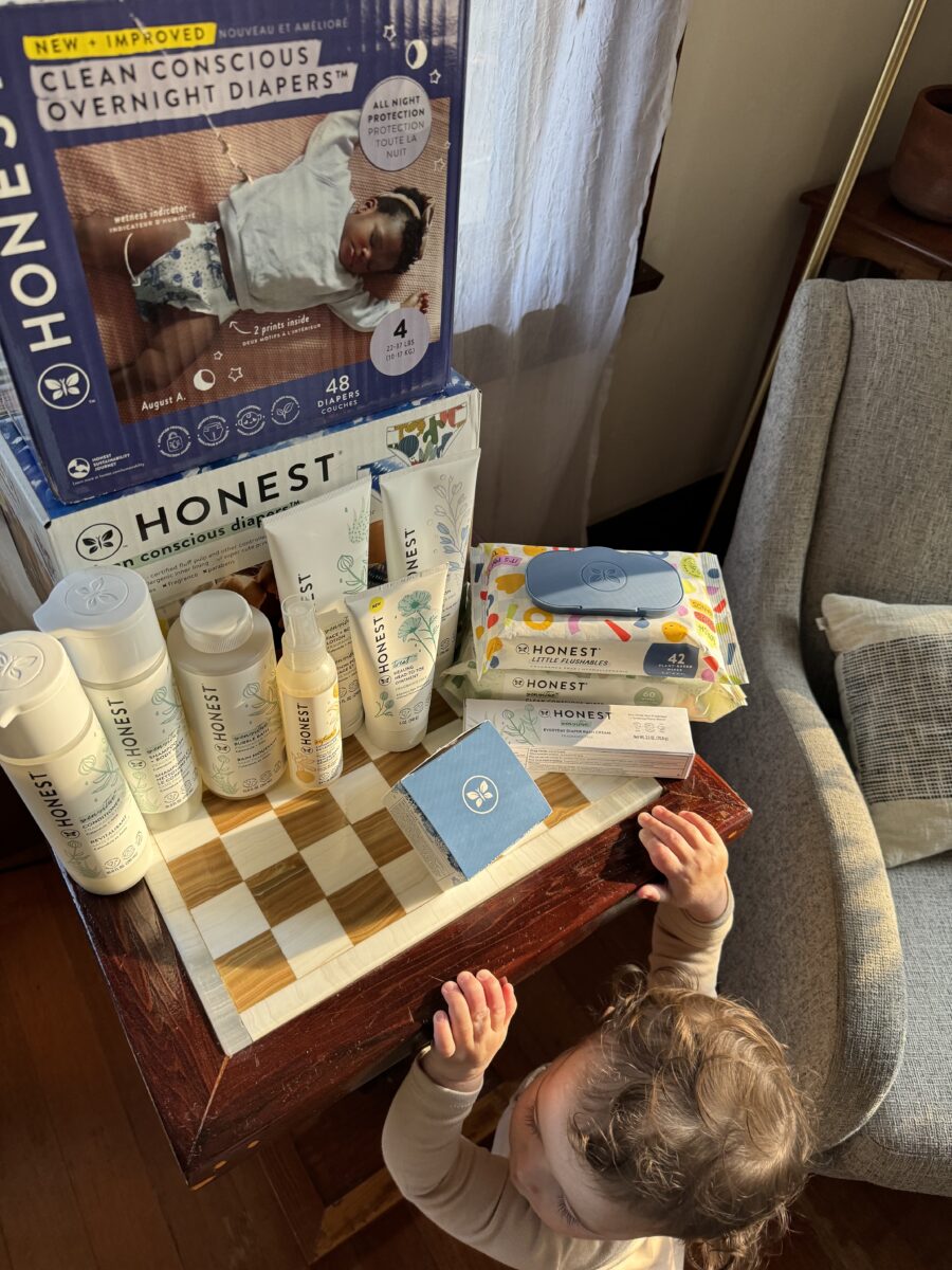 A child reaches up toward a table displaying Honest company diapers, wipes, and baby care products. An armchair is visible beside the table.