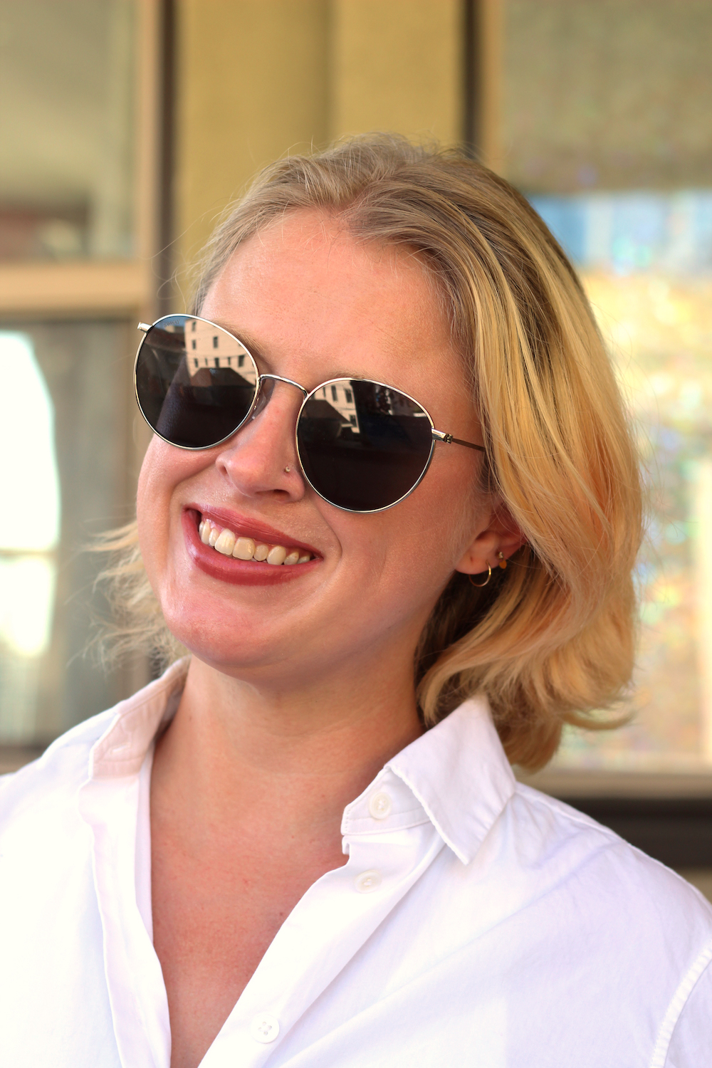 A woman with short blonde hair, wearing sunglasses and a white shirt, smiles in an outdoor setting.