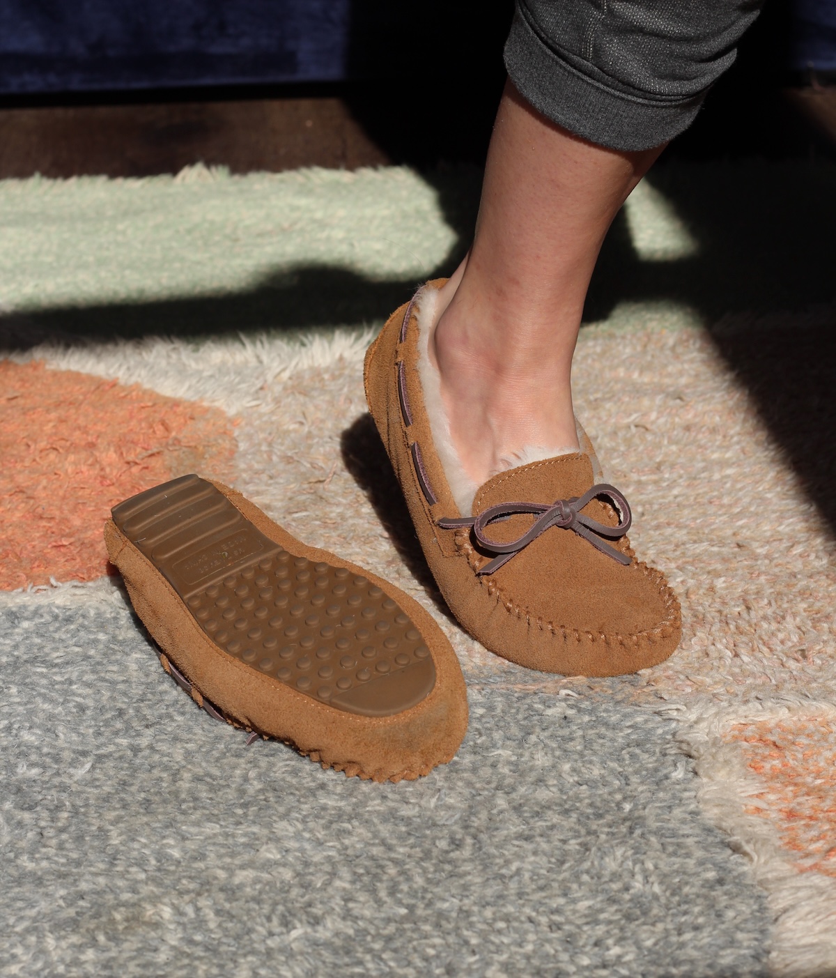 A person wearing one brown moccasin slipper stands on a multicolored rug, next to another slipper placed sole-up.