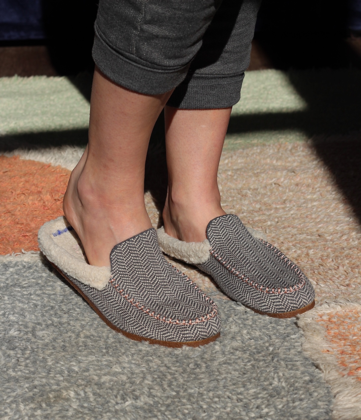 A person wearing gray fabric slippers with white fur lining, standing on a multicolored carpet.