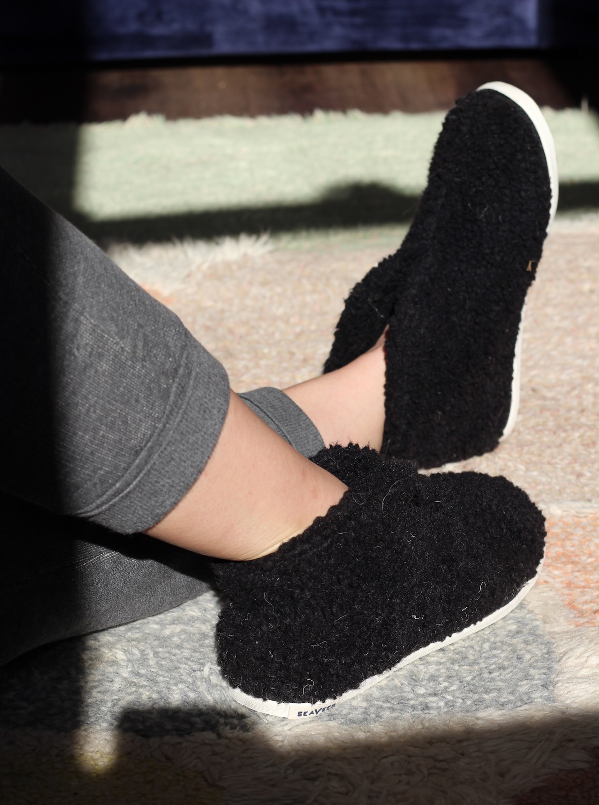 Person wearing fuzzy black slippers and gray pants, sitting on a patterned rug in sunlight.