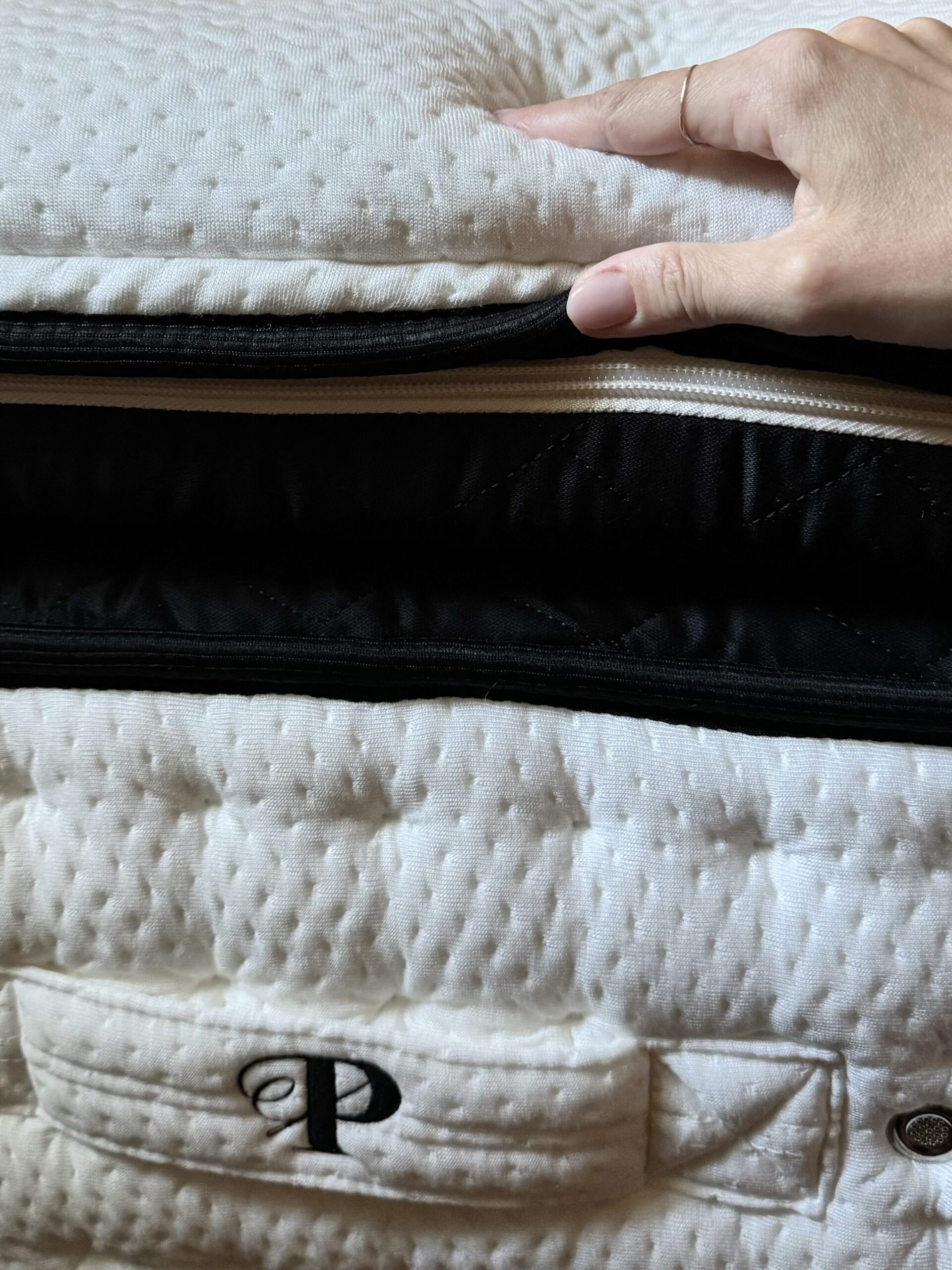 A hand with neatly manicured nails pulls up to reveal the zipped-on latex top of a plush, white, quilted mattress surface.