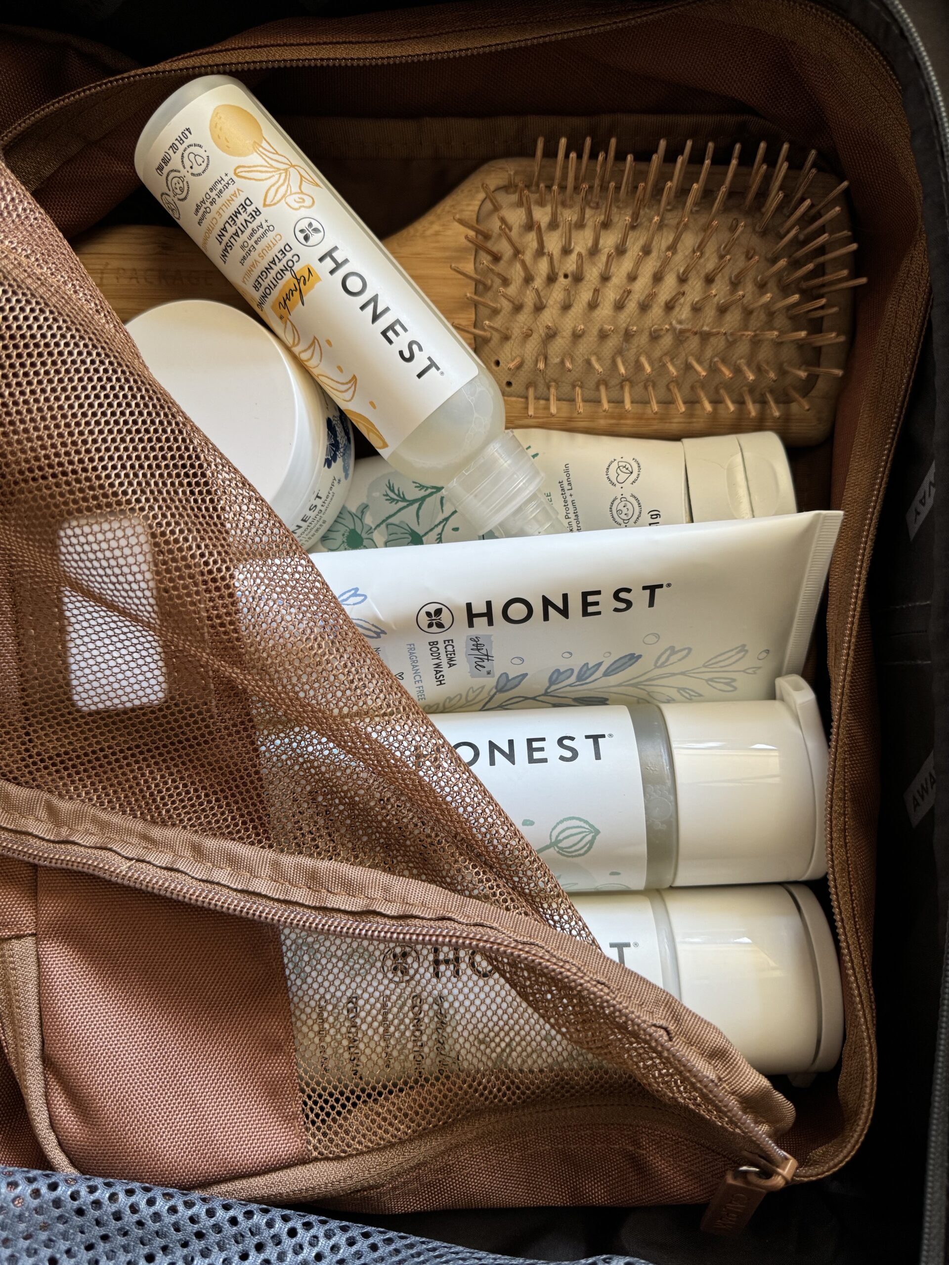 A brown toiletry bag containing various personal care products, including Honest brand bottles, a wooden brush with bristles, and a travel-sized shampoo.