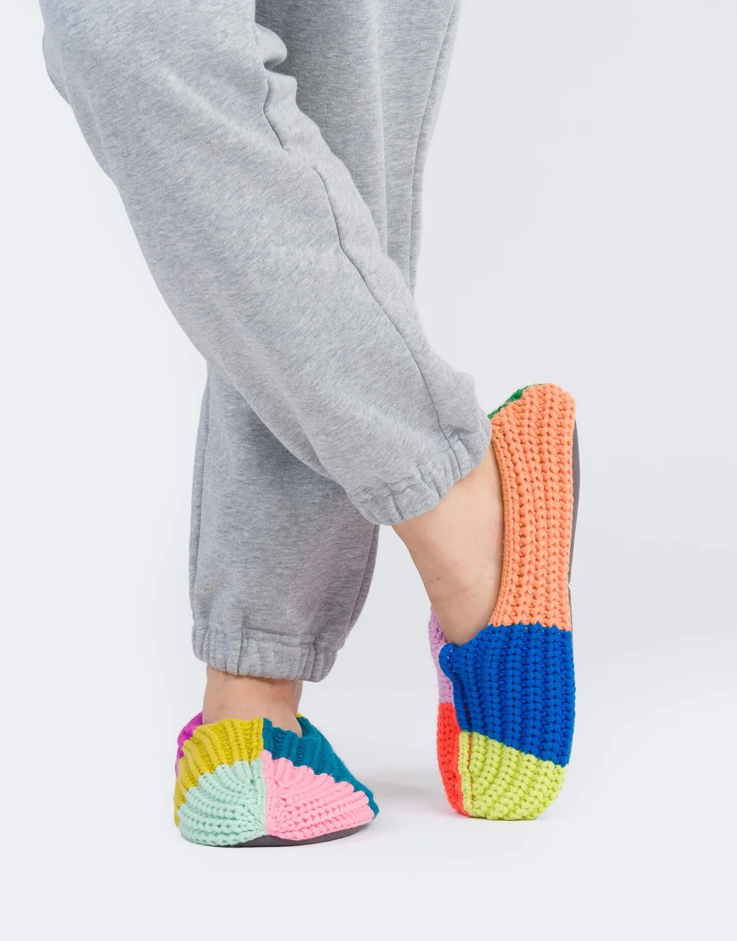 Person wearing gray sweatpants and colorful hand-knitted slippers with a patchwork design, standing against a white background.