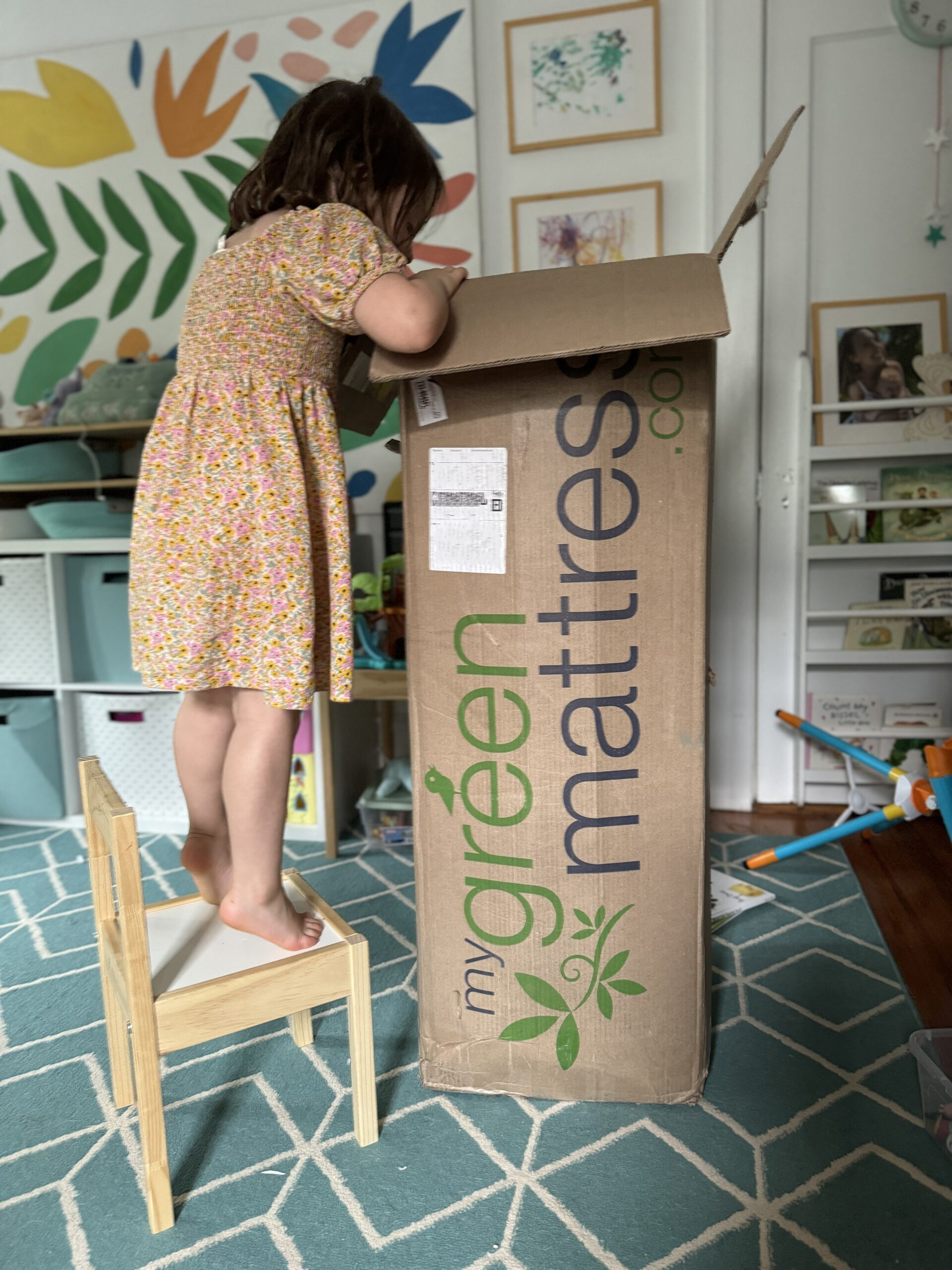 A young girl stands on a chair to look inside a tall cardboard box in a colorful room. The box has "my green mattress" printed on it.