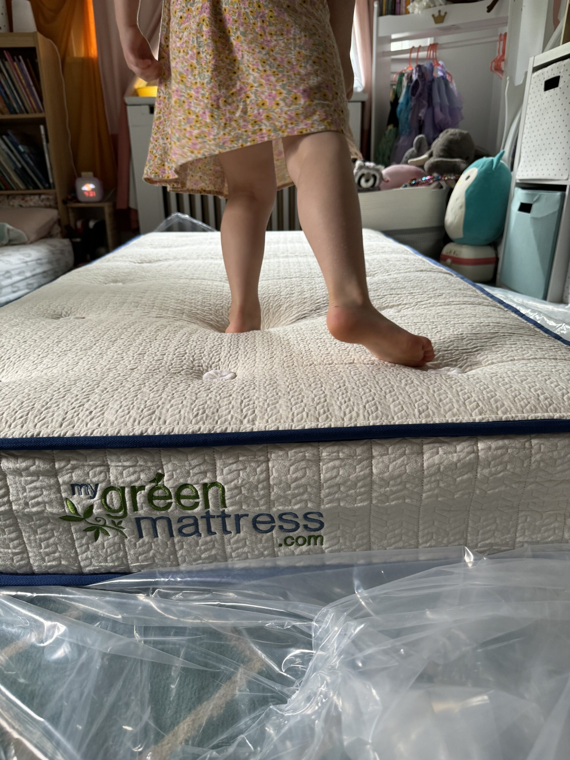 A child stands on a mattress with "green mattress.com" stitched on it in a room filled with various items and furniture.