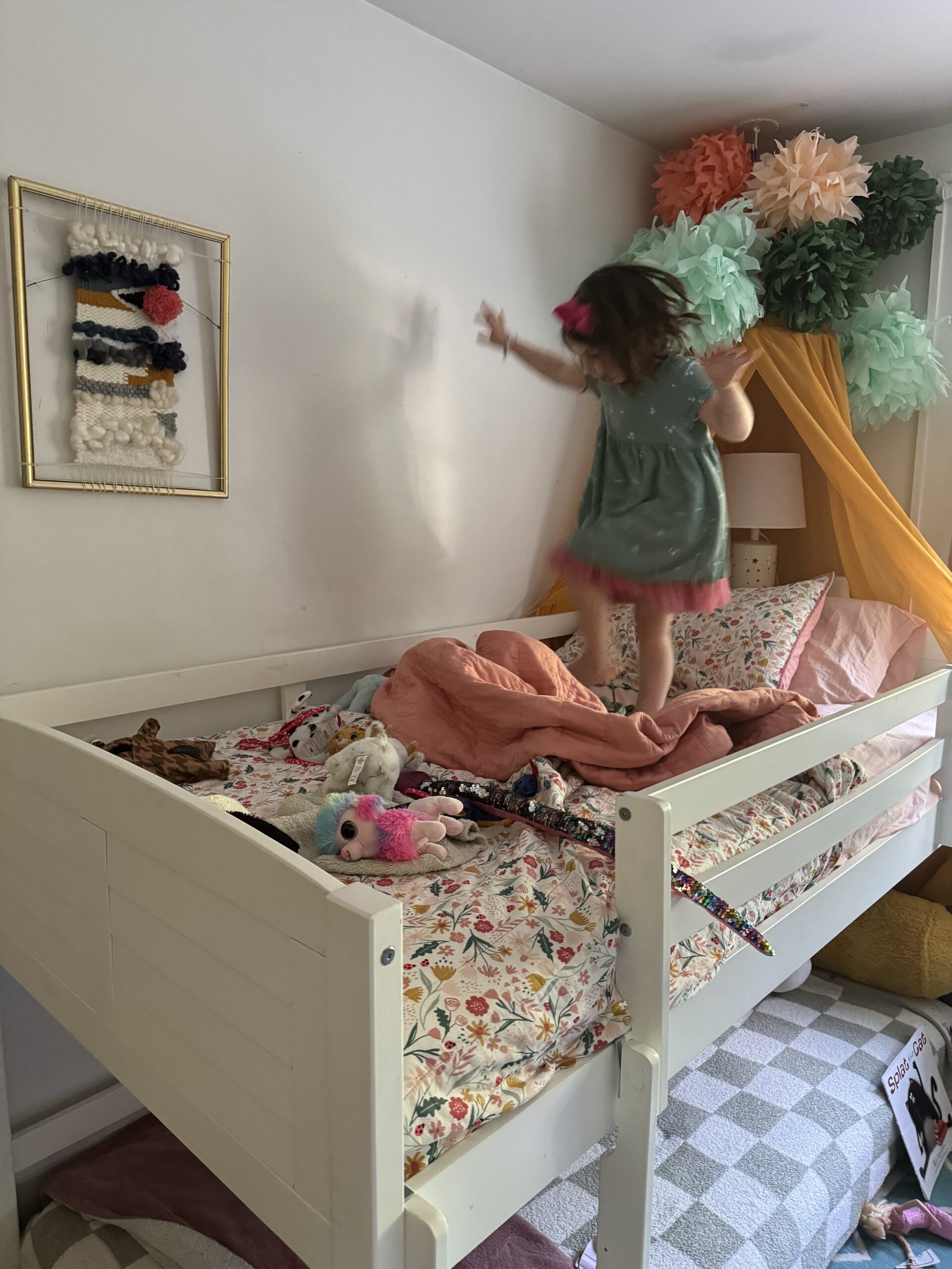 A child in a green dress jumps on a bed with floral bedding, surrounded by toys and pillows. The bed is decorated with a canopy and colorful pom-poms. A woven wall hanging is visible in the background.