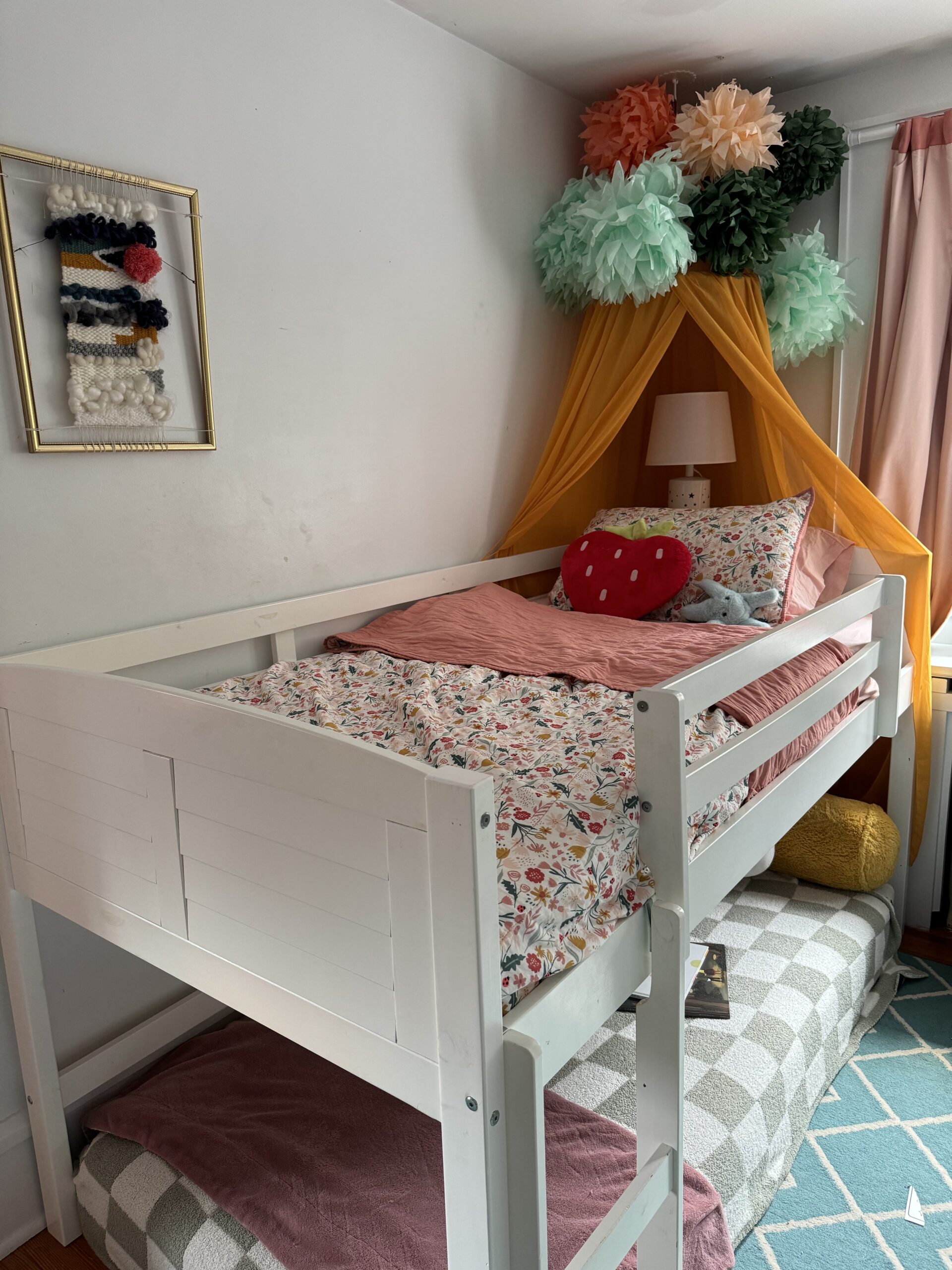 A lofted white bed with floral bedding and pink accents, decorated with hanging paper pom-poms and an orange canopy. A small lamp and a knitted wall hanging are visible in the background.