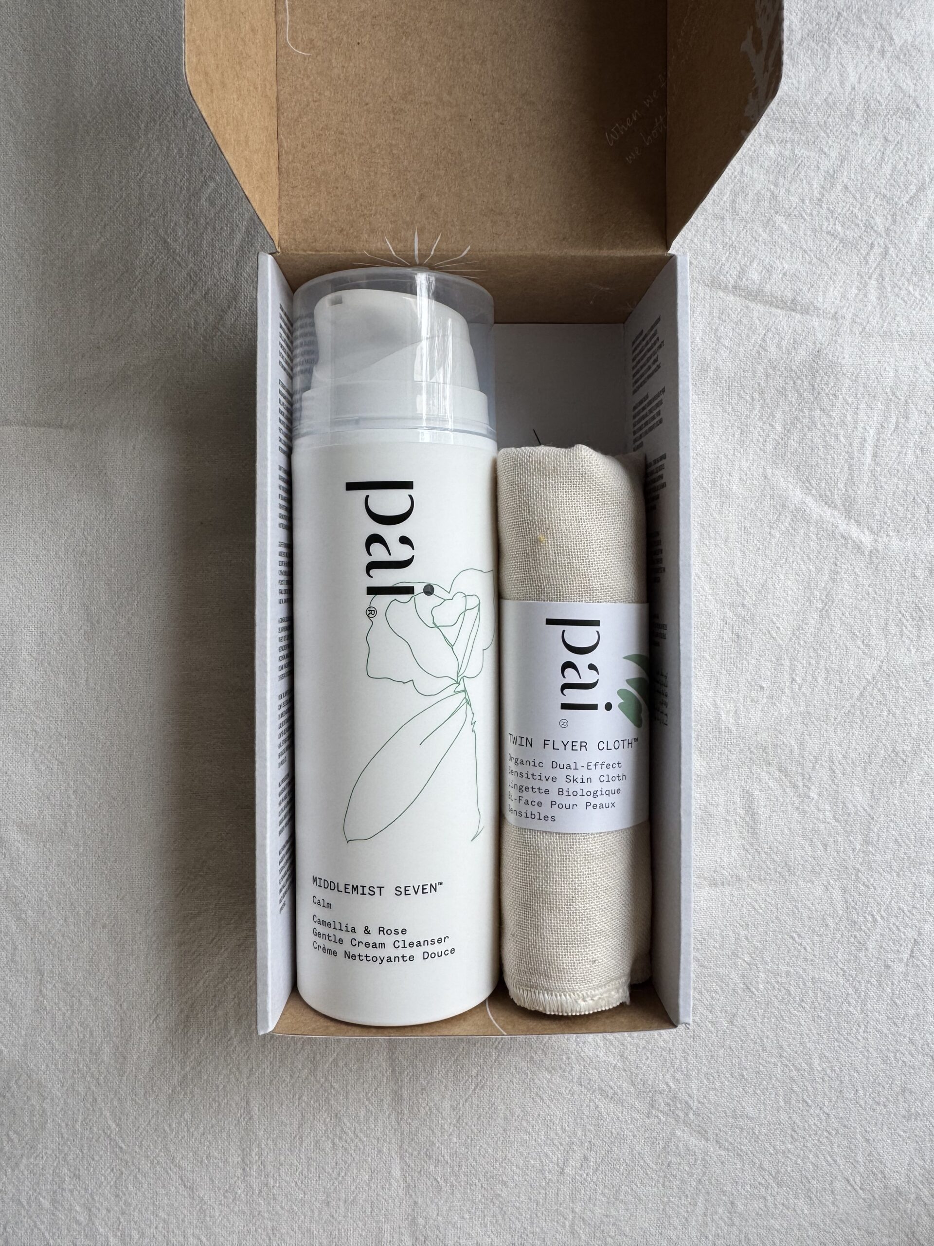 A bottle of Pai face cleanser and a towel inside a cardboard box on a white fabric surface. 