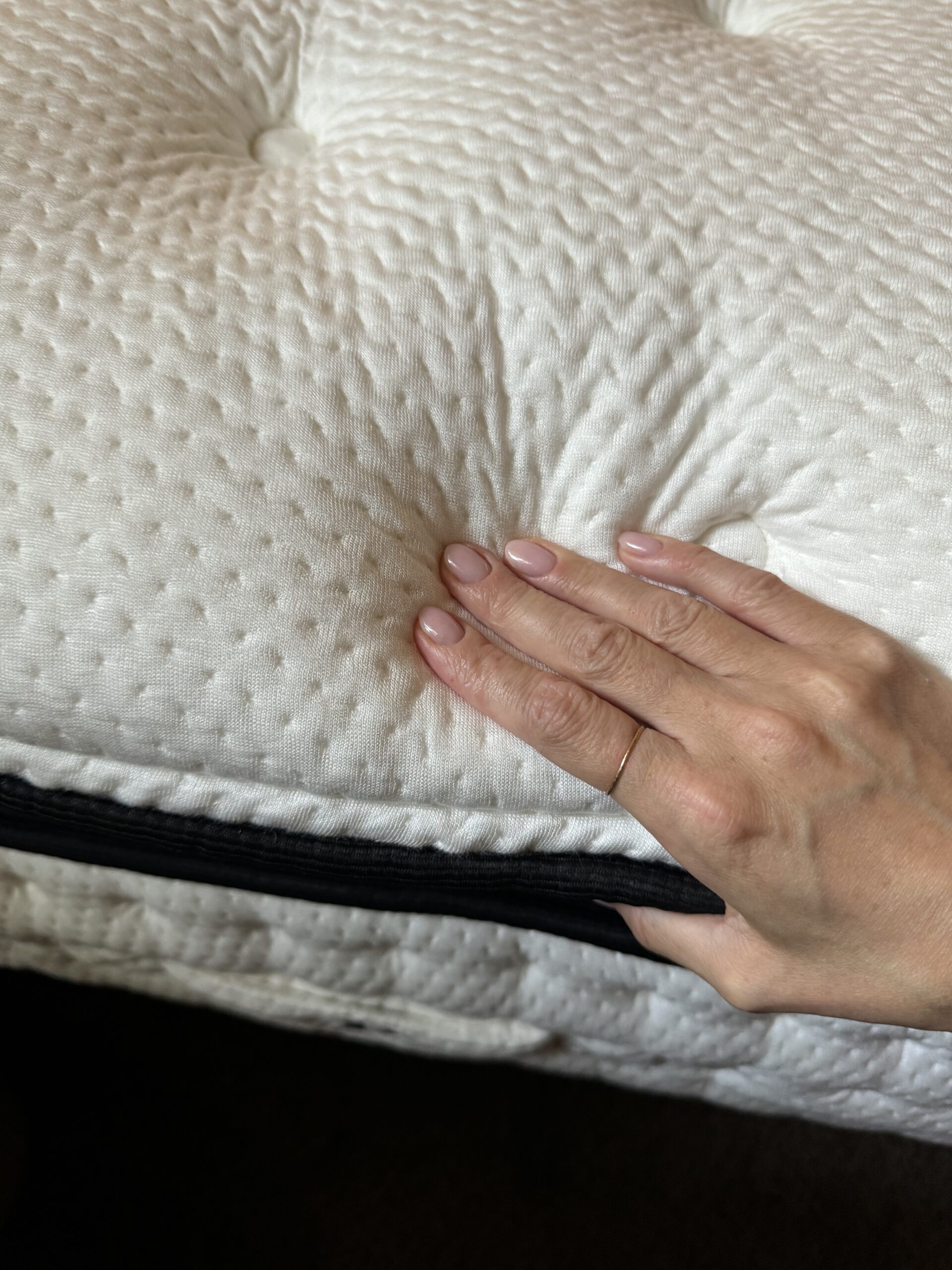 A hand with pale pink nails presses down on a thick, white, tufted quilted surface of an organic mattress.