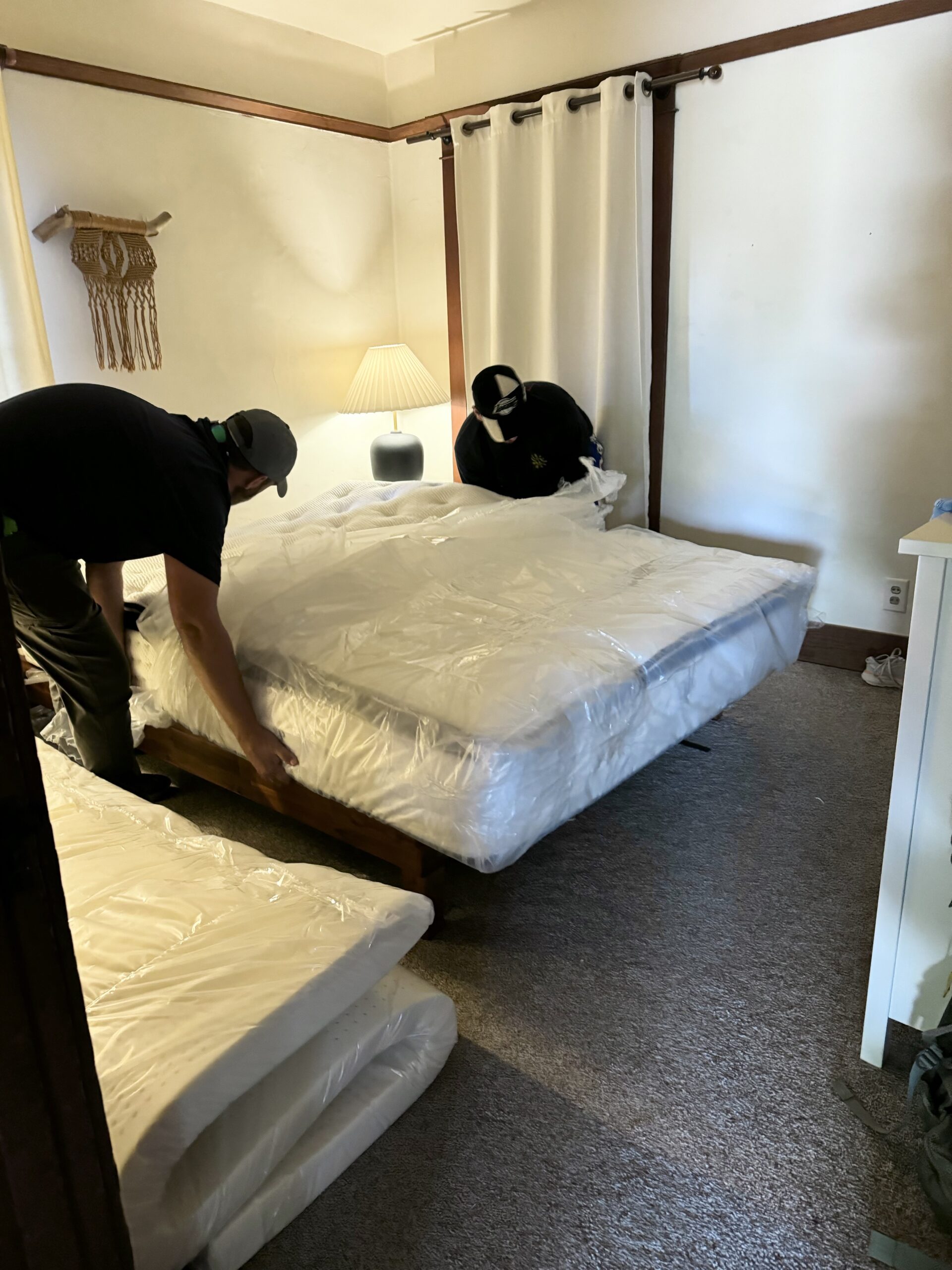 Two people are placing a mattress onto a bed frame in a room with beige carpet, white walls, and a lamp on a side table.