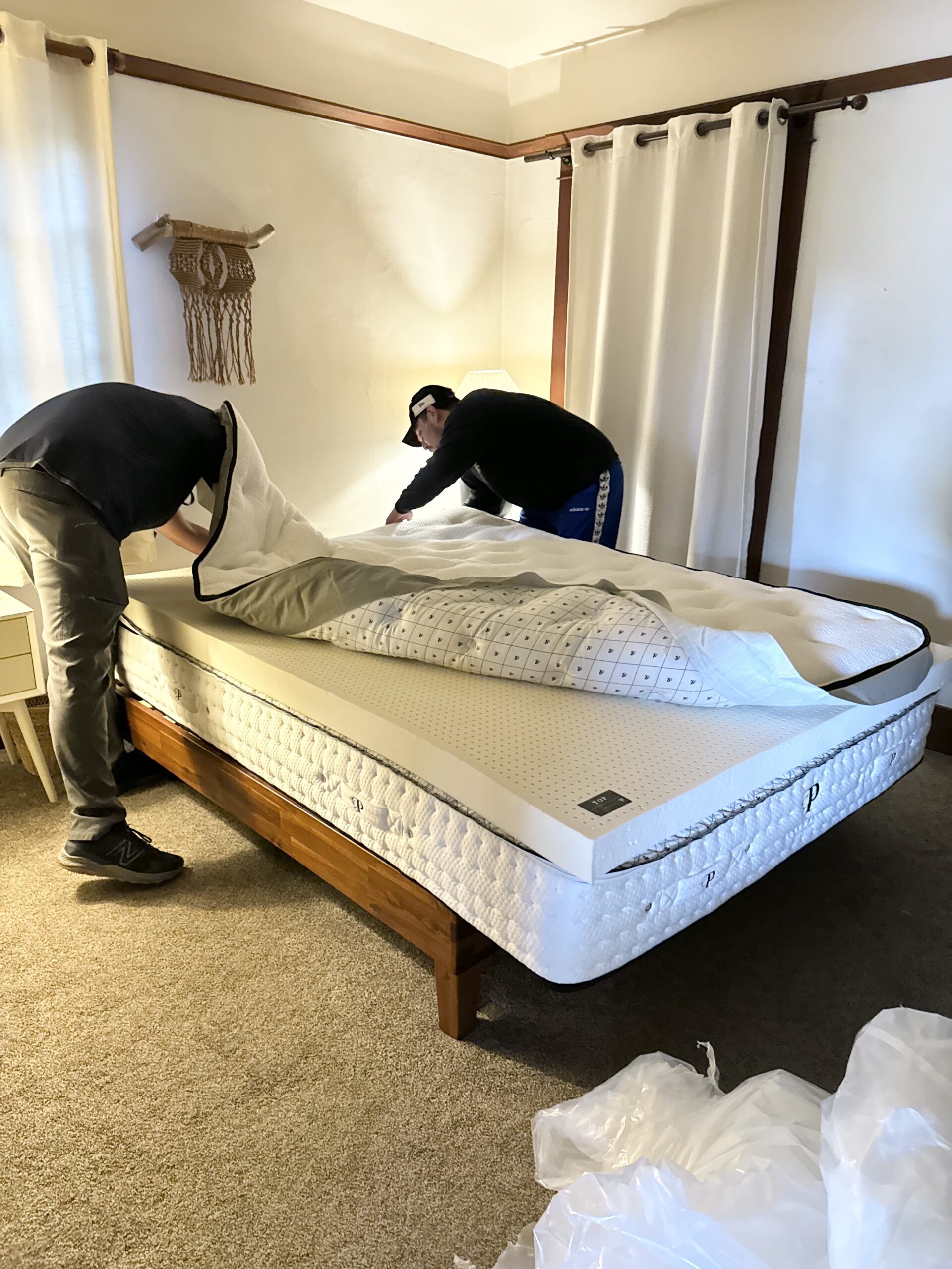 Two people are making a bed with a white mattress on a wooden frame, adjusting a mattress cover in a well-lit room.