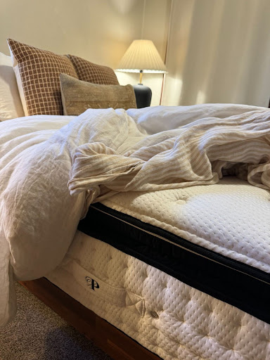 A bed with white linens, pillows, and a beige checked throw pillow. The linens appear slightly disheveled. A bedside table with a lamp is visible in the background.