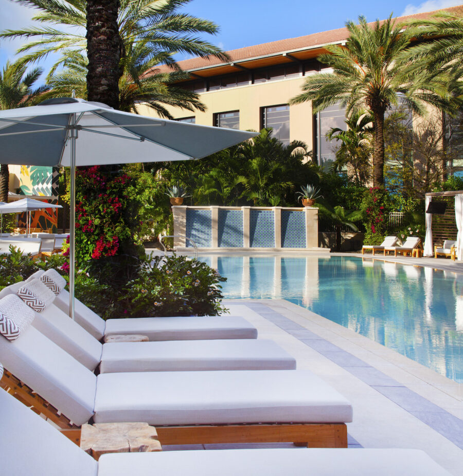 Poolside lounge chairs with white cushions are shaded by umbrellas, surrounded by palm trees and lush plants. A modern building is in the background.