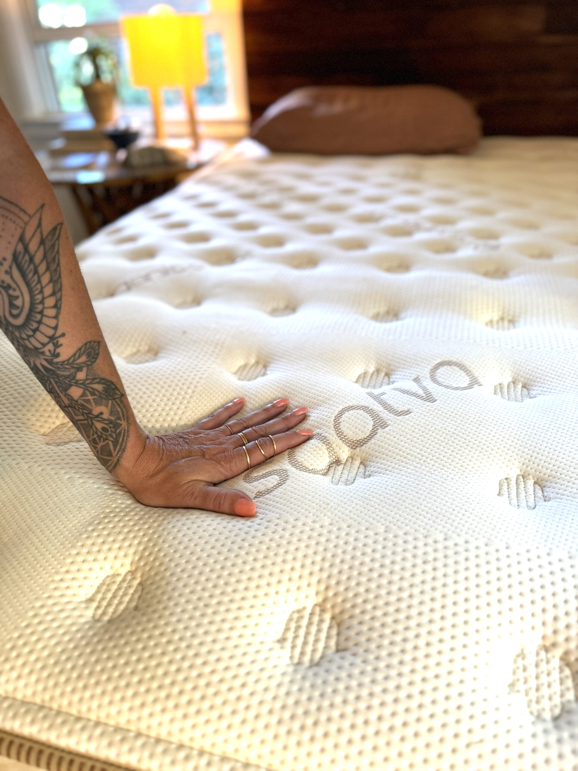 A hand with rings and a tattoo presses down on a Saatva mattress, showing its texture and indentation. A bedside table with a lamp and cushion are visible in the background.