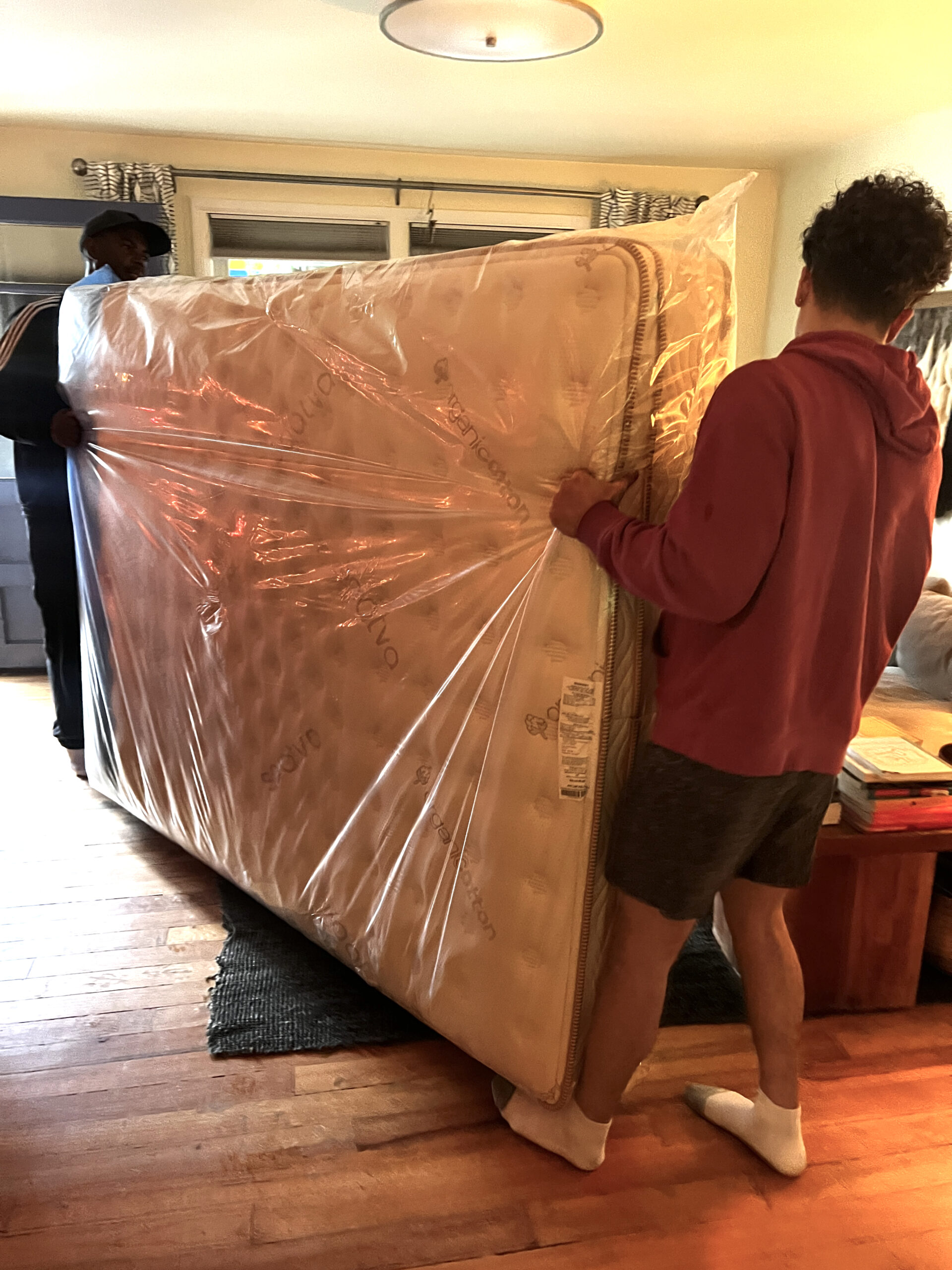 Two individuals are moving a mattress wrapped in plastic in a living room.