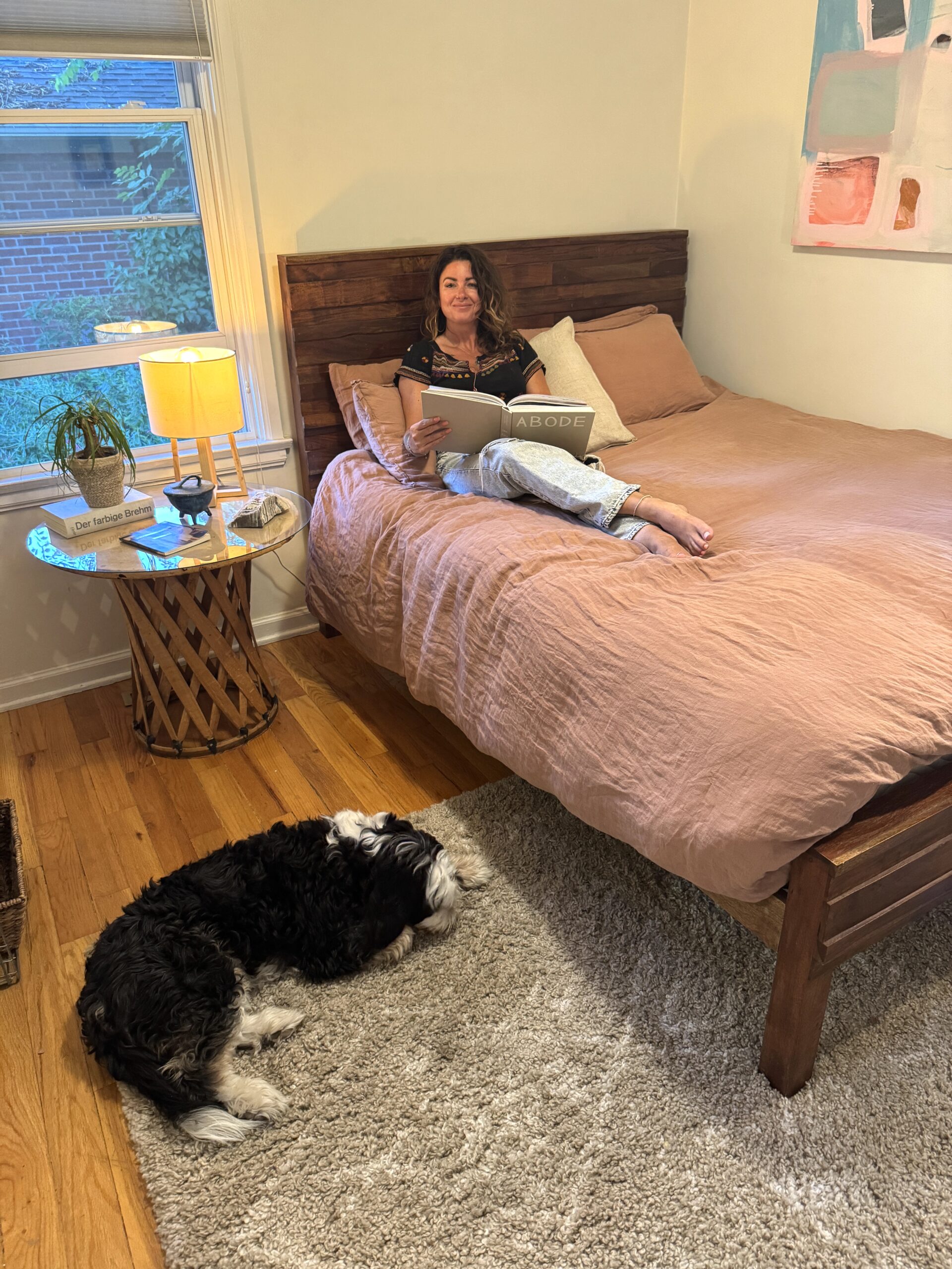 A woman sits, reading, on top of a wooden bed frame covered in pink linens. There is a light brown side table with a lamp, and a potted plant. A painting hangs on the wall above the bed, and a window with blinds is on the left. There is a fluffy black and white dog lying on the floor.