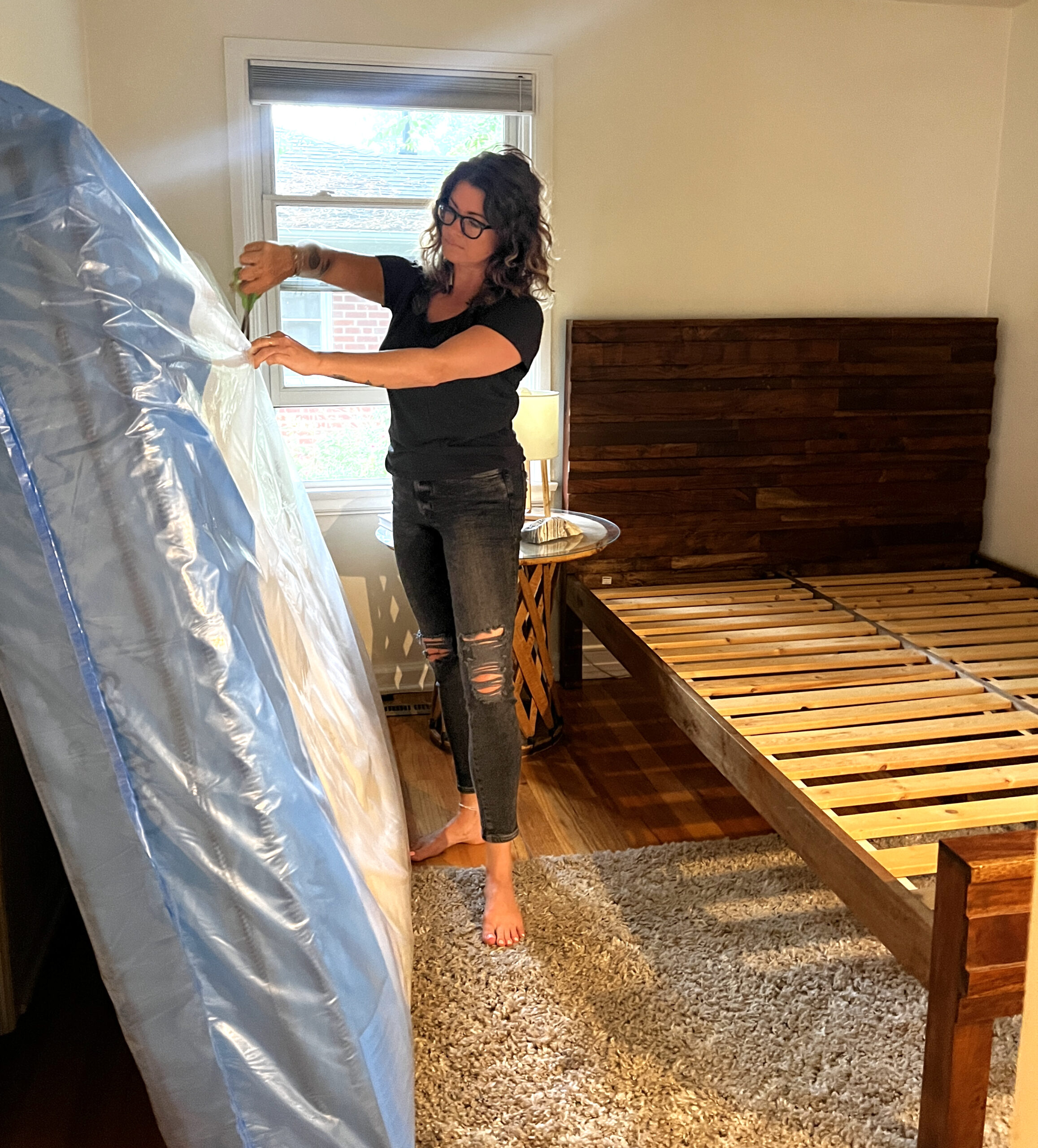 A person with glasses and dark hair is standing barefoot while removing plastic from a mattress. The room has a wooden bed frame and a beige rug. A window with blinds is in the background.