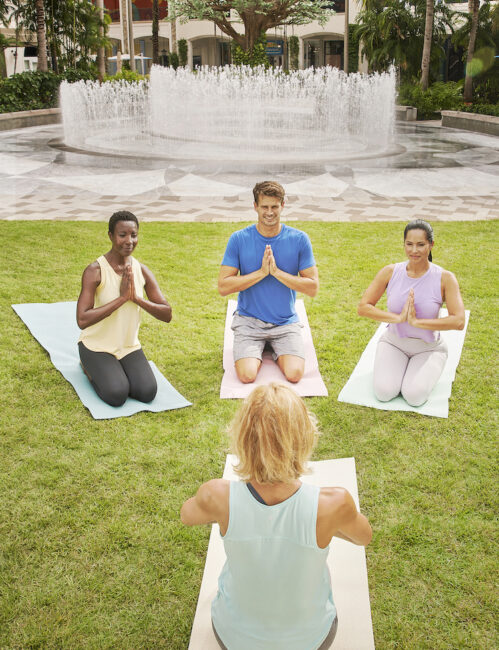 Four people are kneeling on yoga mats in a grassy area, performing a yoga pose with their hands in prayer position. A large fountain is visible in the background.