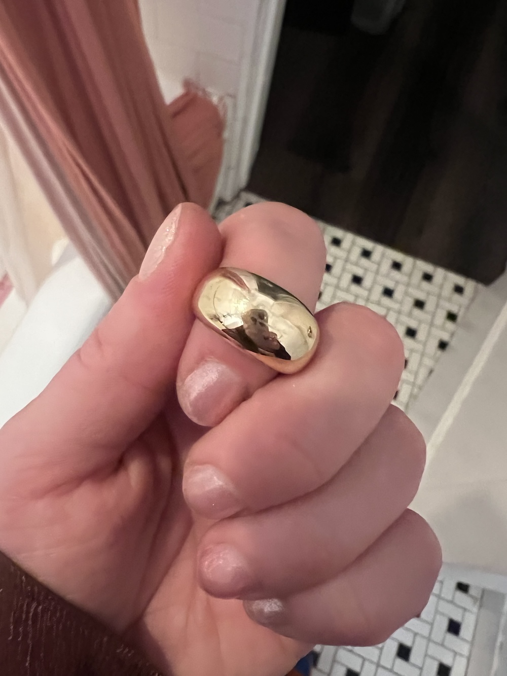 A hand holding a shiny gold ring with a reflection of the person taking the picture. Background includes a tiled floor and part of a curtain.