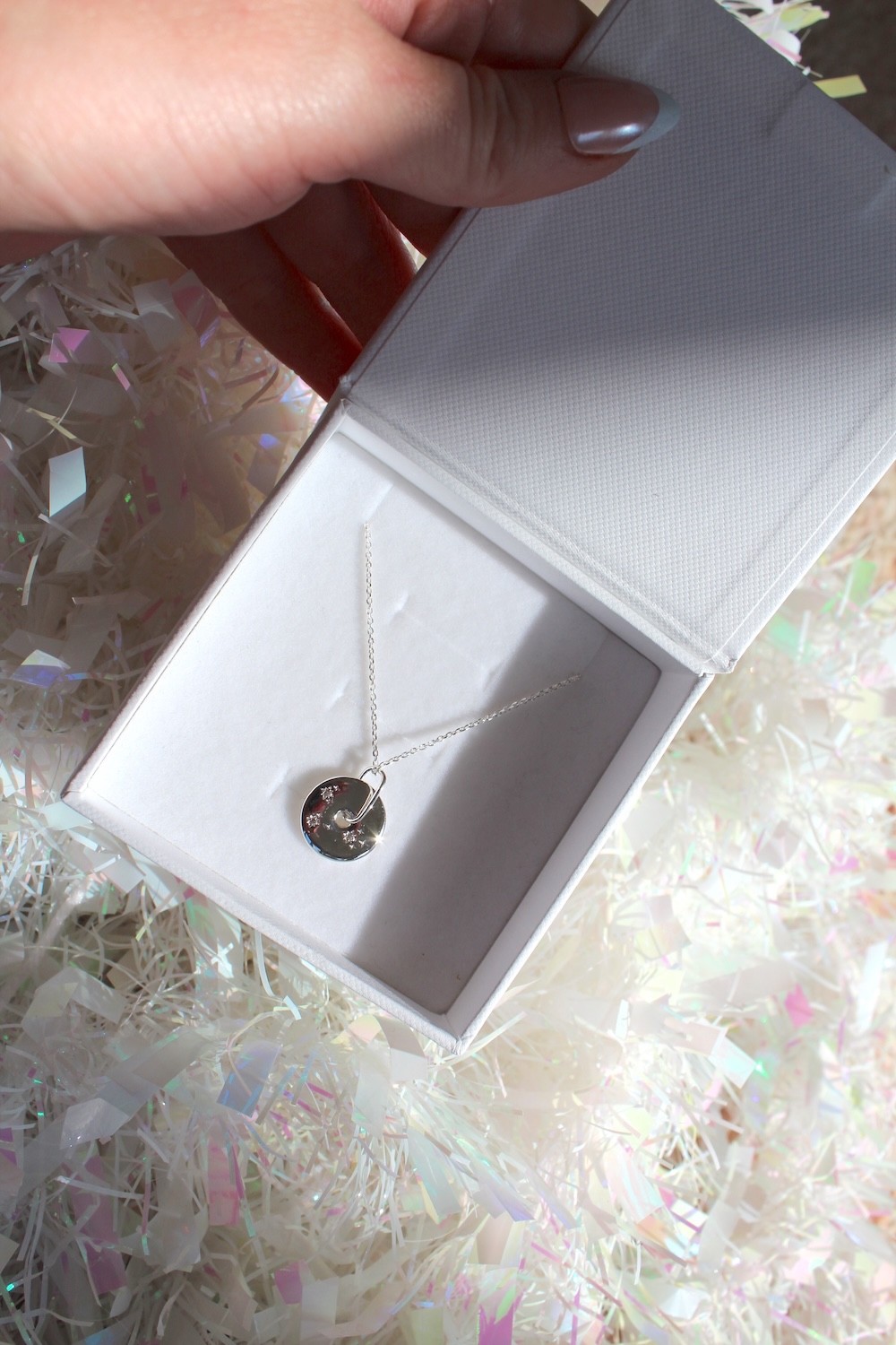 A hand holds an open white box containing a silver necklace with a round pendant, surrounded by iridescent paper.