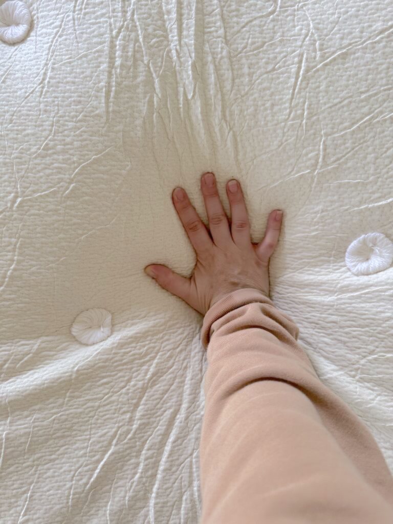 A hand pressing down on a white, textured organic mattress, causing an indentation. The person is wearing a beige sleeve.