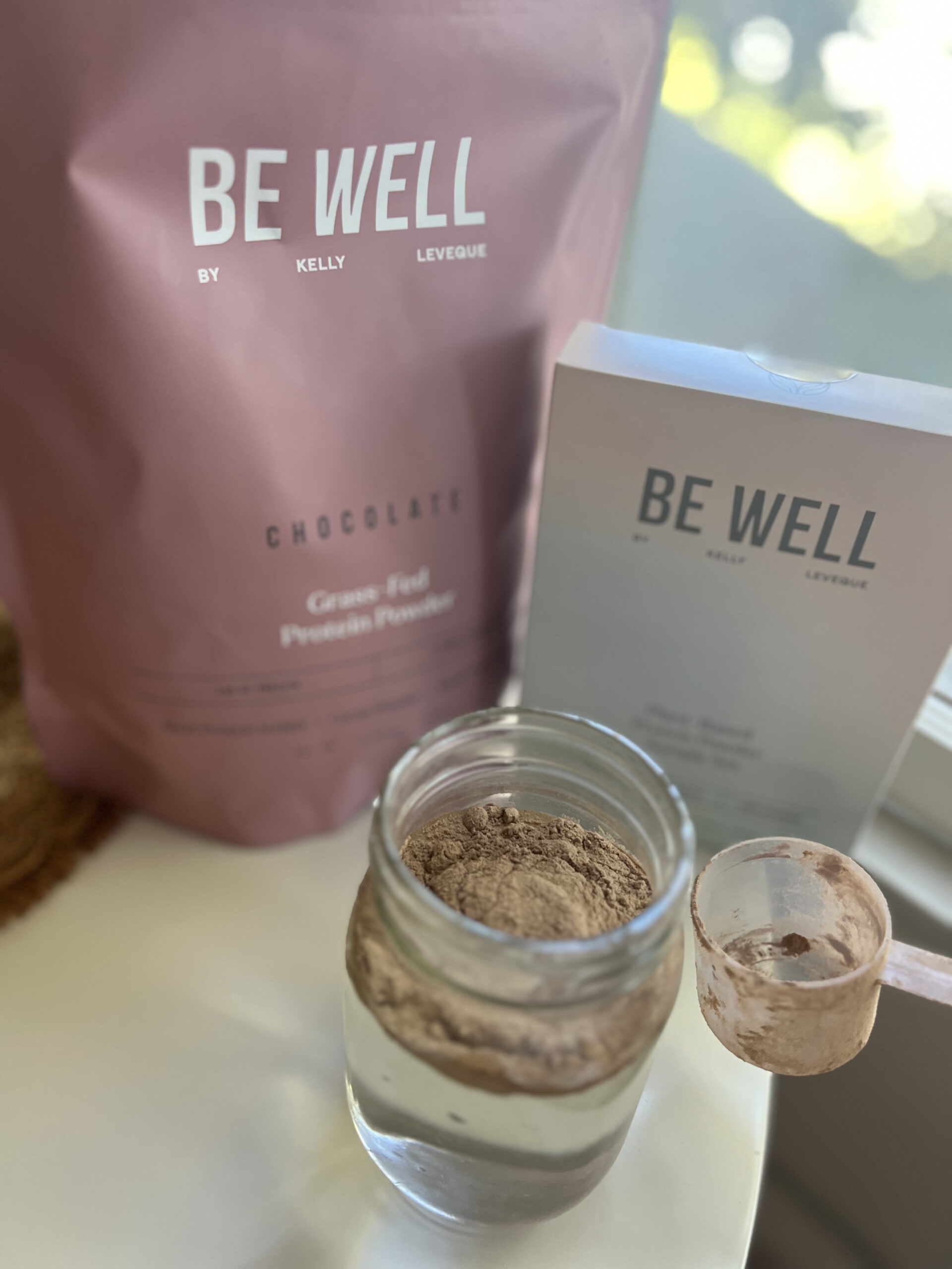 A jar of chocolate protein powder beside a branded pouch and box labeled "Be Well." A scoop rests on the jar's rim.