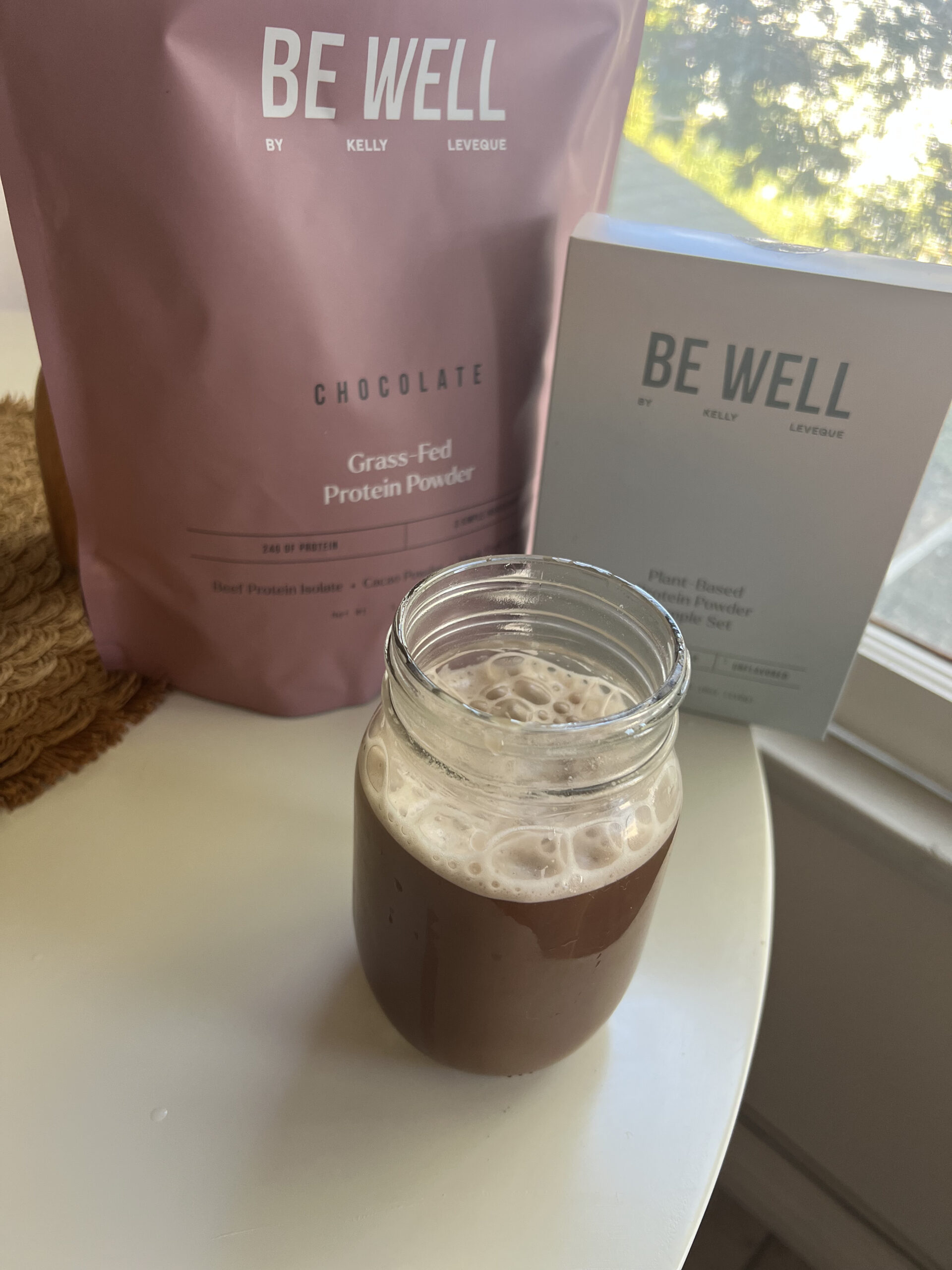 A glass mug filled with a chocolate protein shake is next to a pink bag of chocolate grass-fed protein powder and a white box on a table by a window.