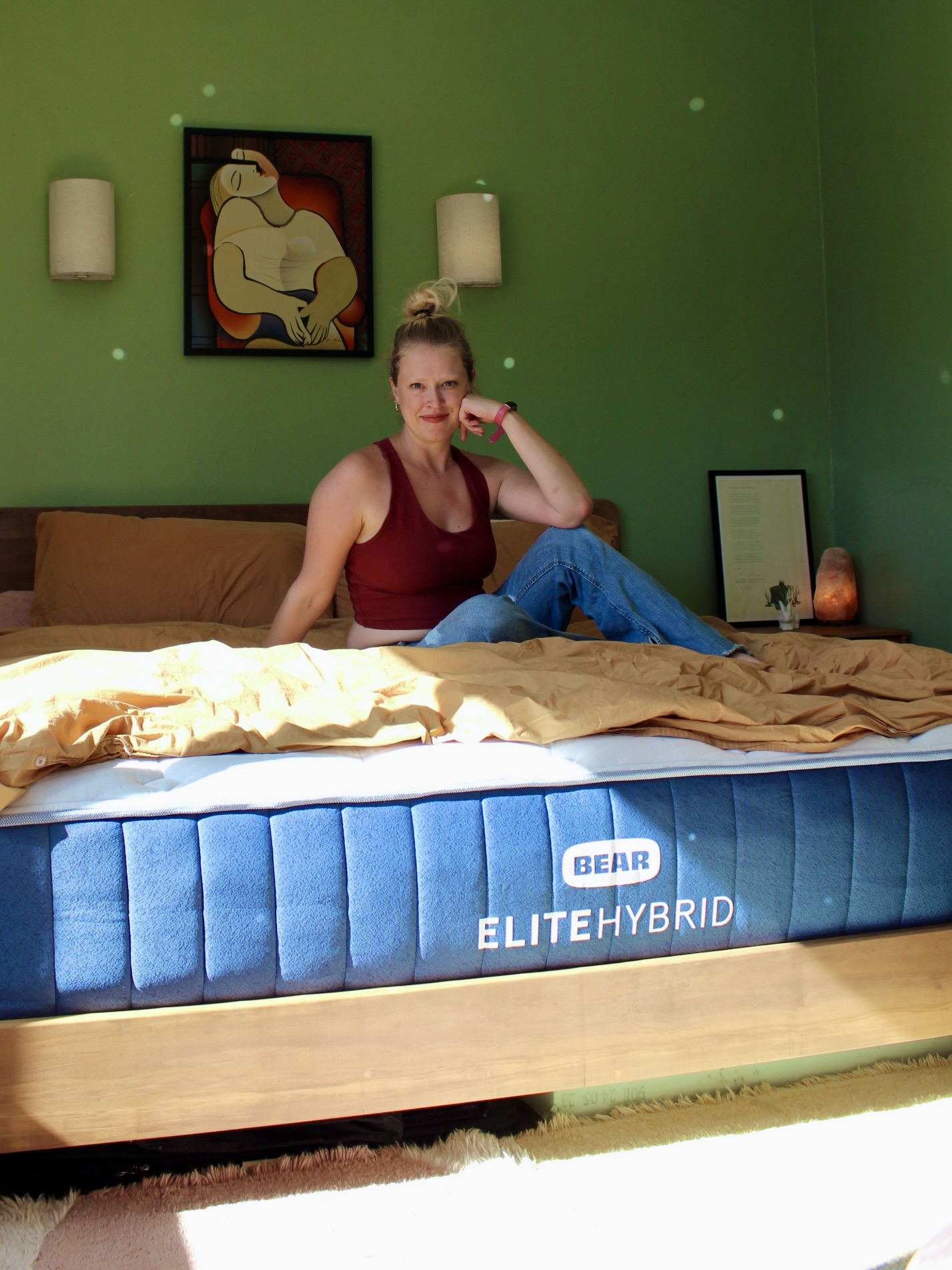 A person sits on a bed with a blue mattress labeled "Bear Elite Hybrid" in a green-walled room with a framed artwork and two sconces.