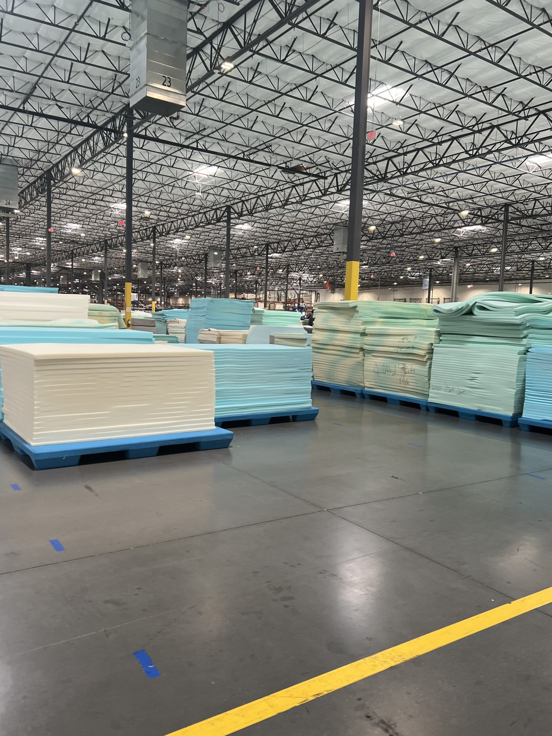 Large stacks of foam sheets on blue pallets in a spacious warehouse with high ceilings and overhead lights.