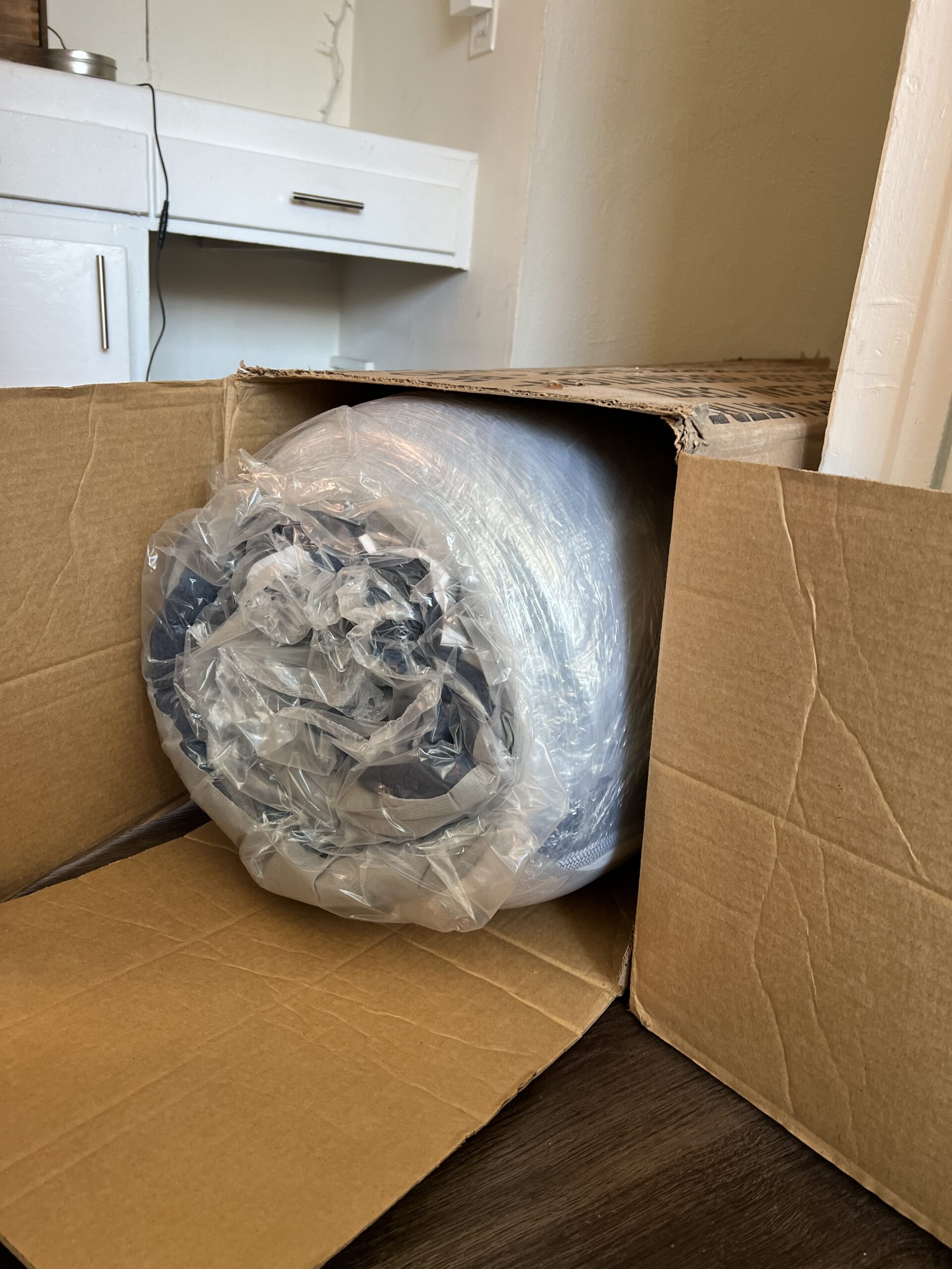 A rolled up mattress is partially unwrapped from plastic packaging inside an open cardboard box, placed on the floor next to a white cabinet.