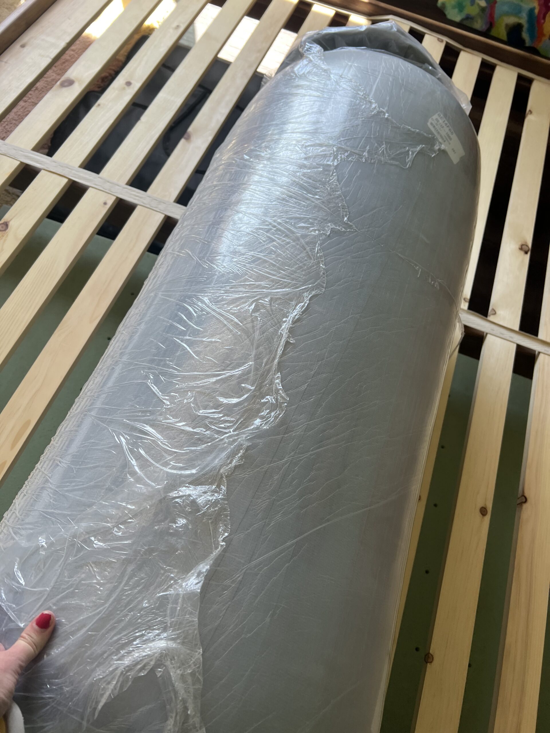 A rolled-up mattress in plastic wrapping is placed on a wooden slatted bed frame.