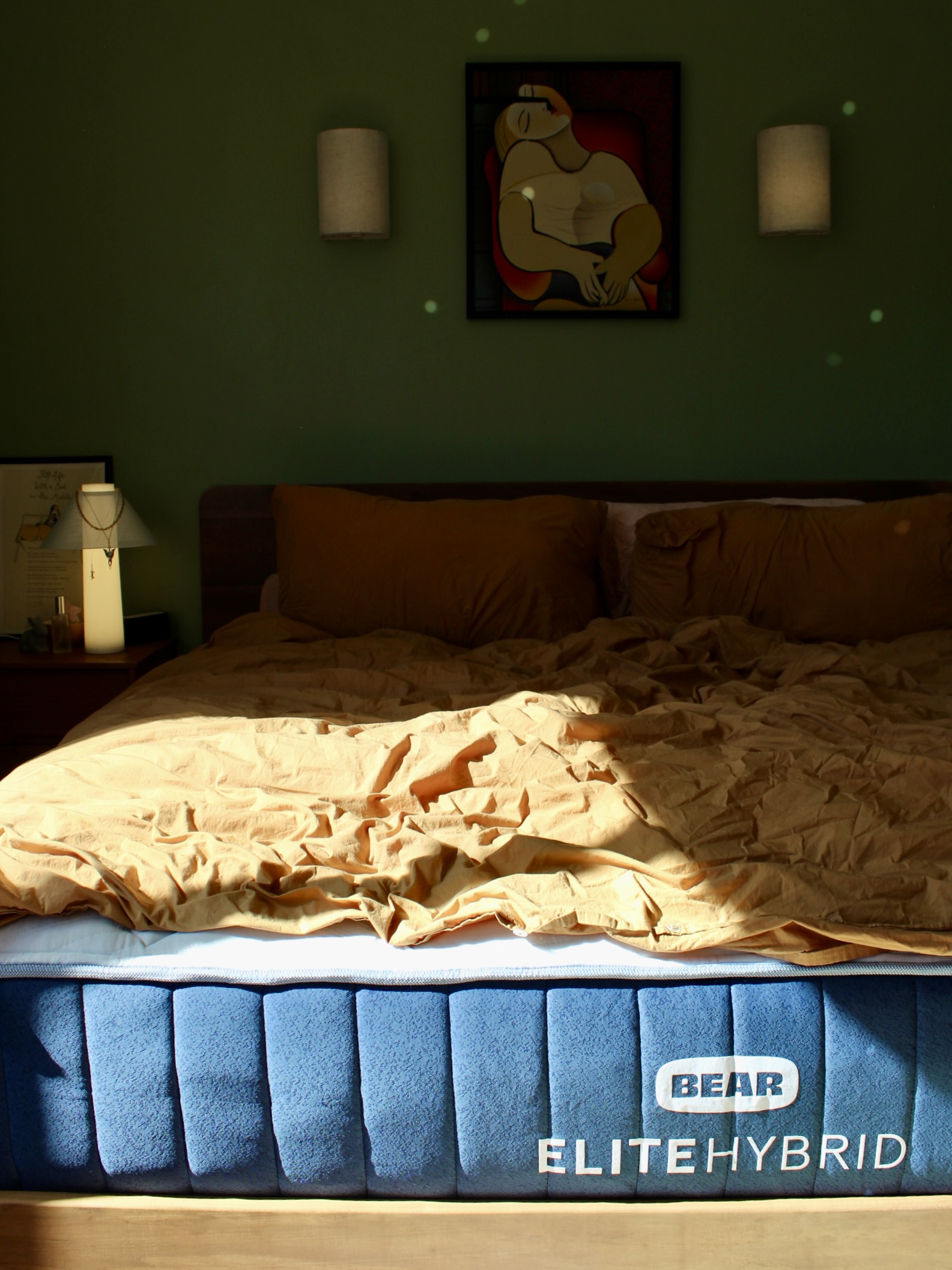 A sunlit bedroom with a blue Bear Elite Hybrid mattress, crumpled tan bedding, two wall lamps, and a framed artwork on the green wall.