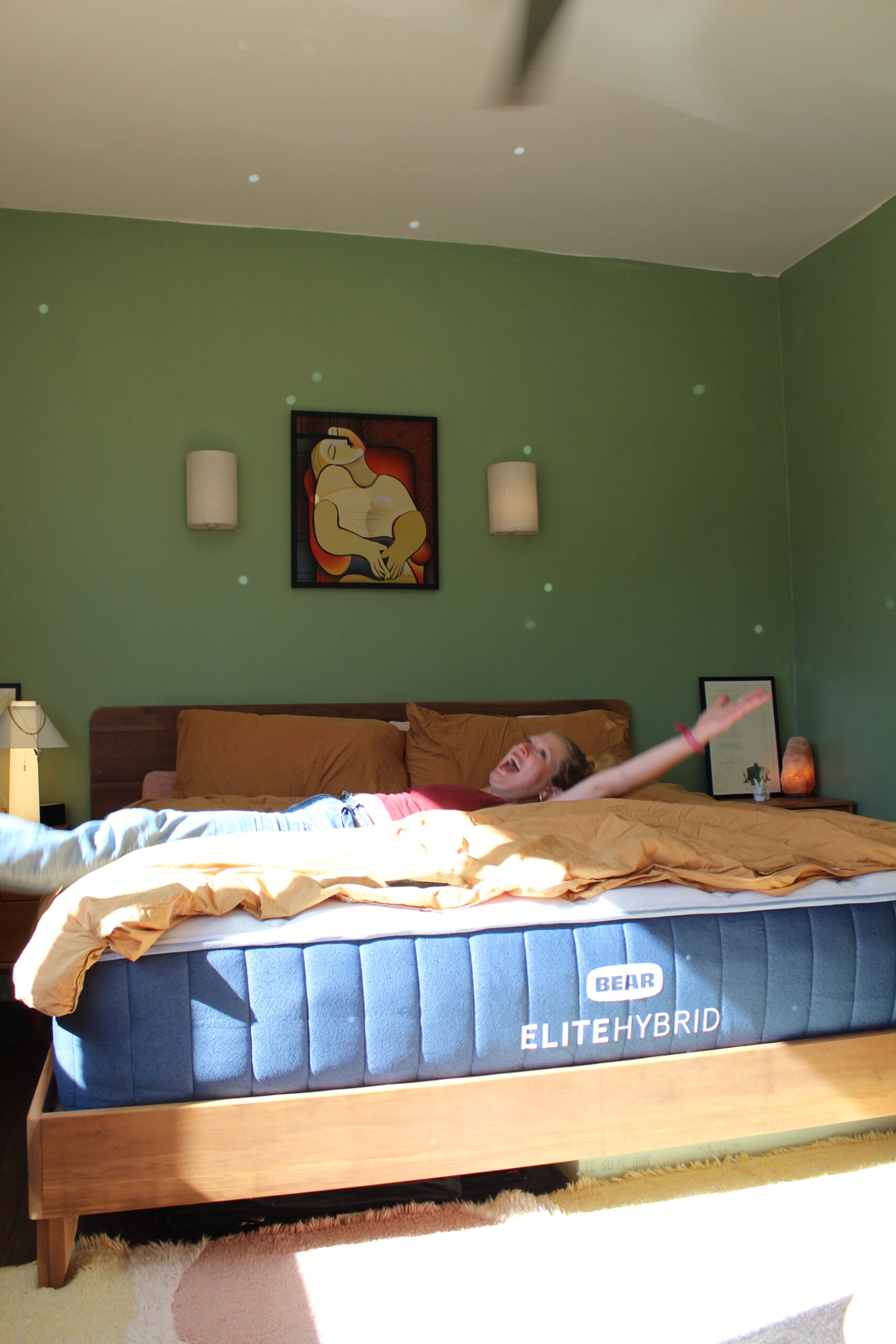 A person is lying on a bed with arms outstretched, in a room with green walls, wall art, and a Bear Elite Hybrid mattress. Light spots are visible on the walls.