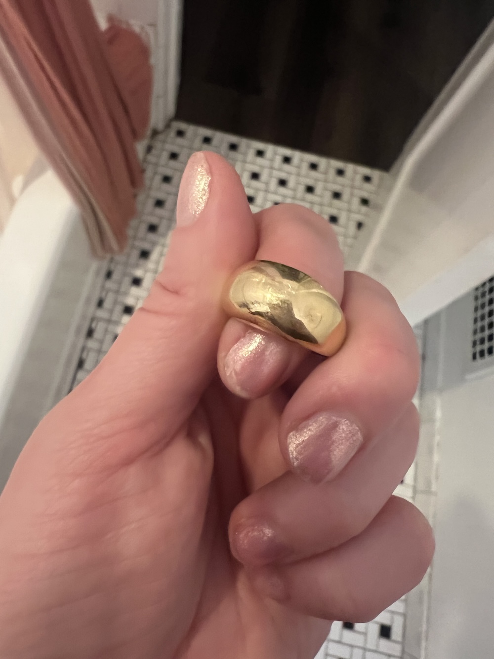 Hand holding a gold ring with an engraving. Background shows a tiled floor and a towel.