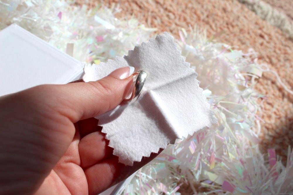 A hand holding a silver ring with a white cloth for polishing. White and pink decorations are in the background.