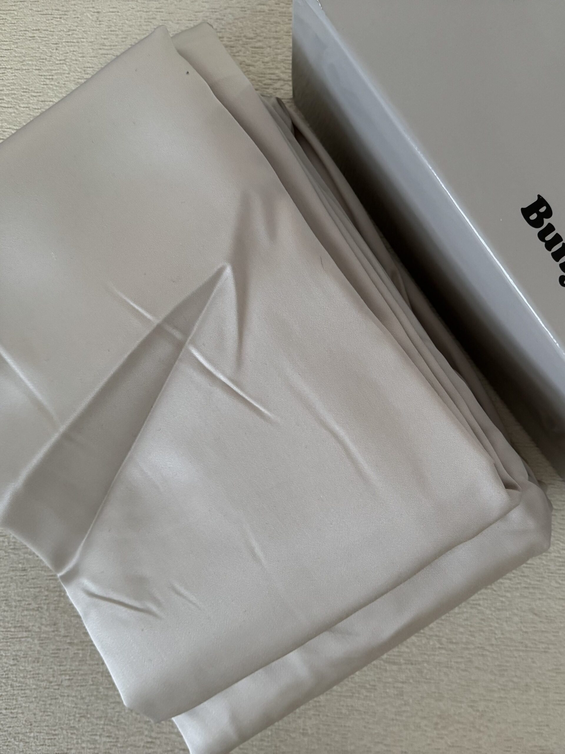 A folded beige bedsheet next to a gray box labeled "Buffy" on a beige surface.